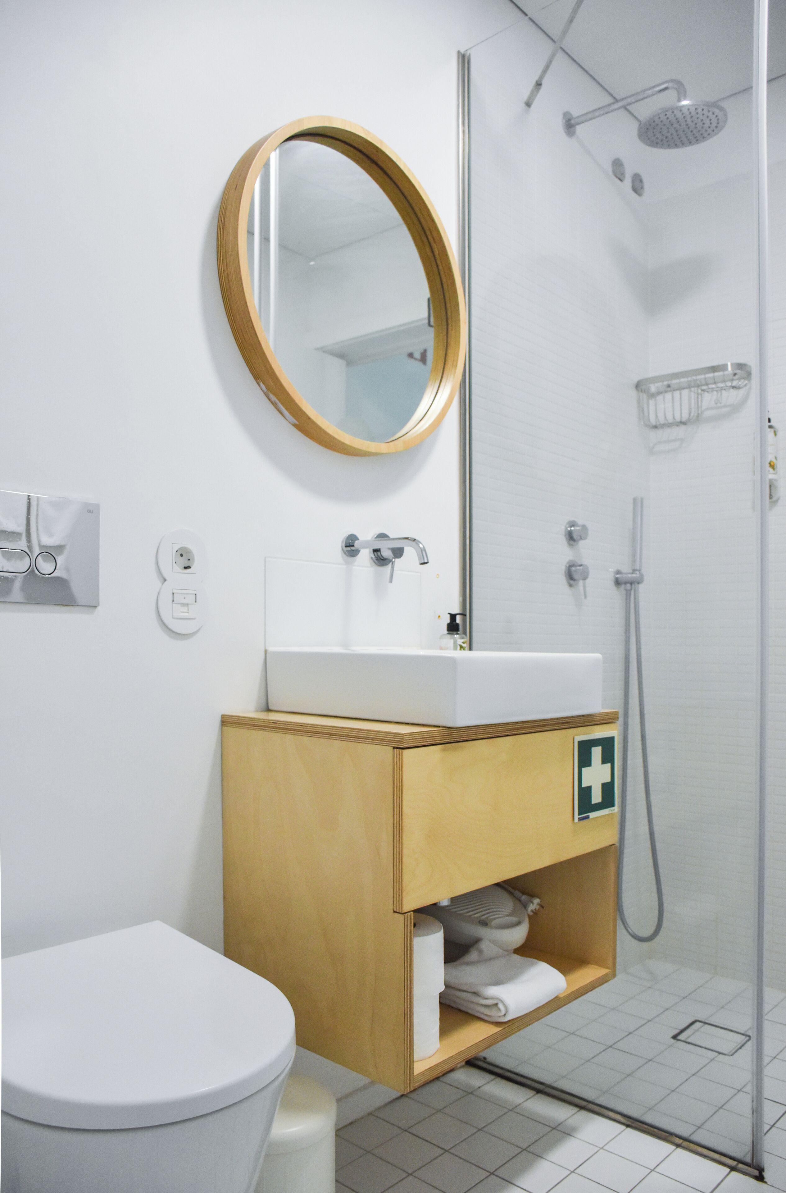 Et badeværelse med et hvidt toilet, et træskab med en vask, et spejl og en bruseniche.