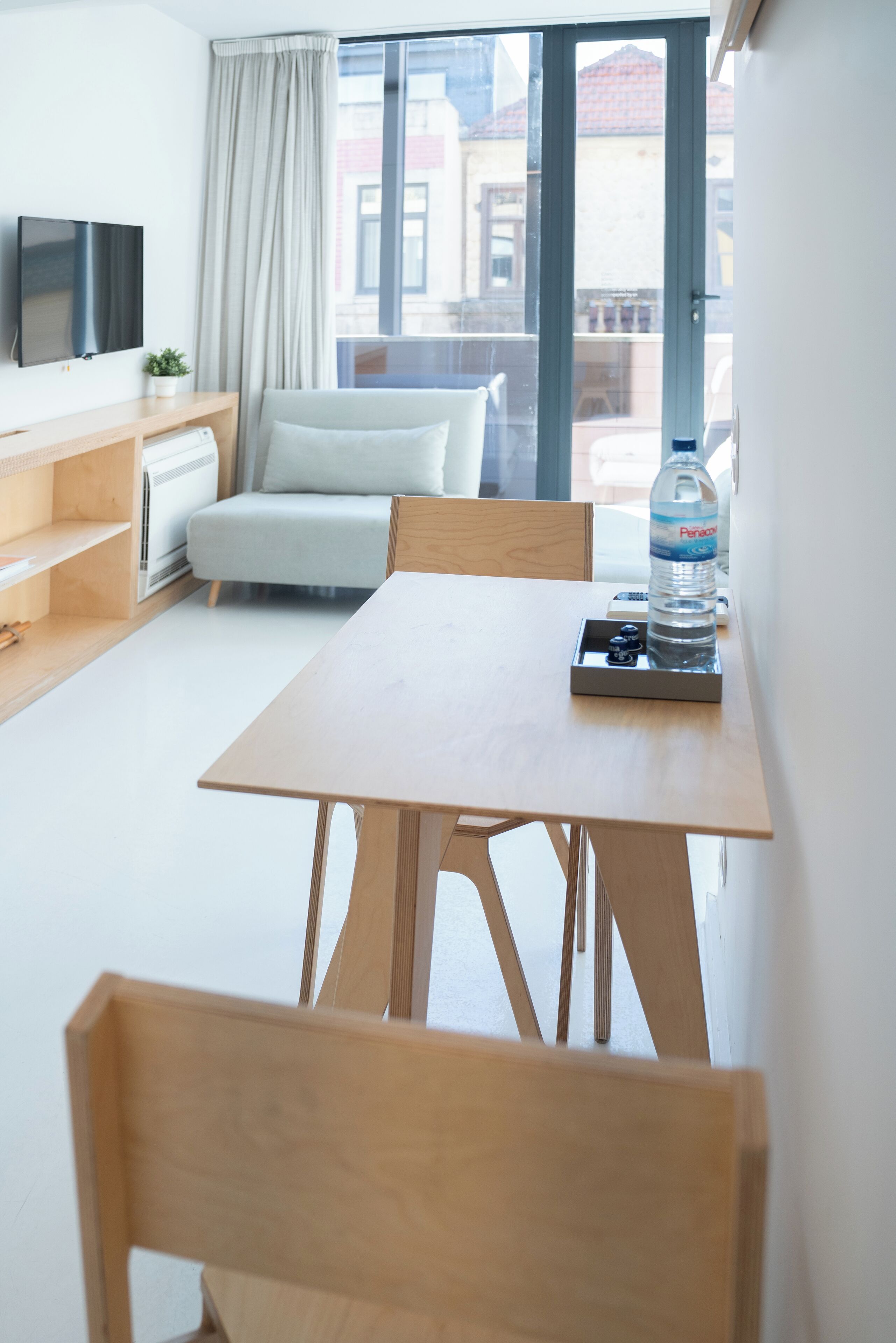 Een moderne hotelkamer met een houten tafel, een sofa, een televisie en een raam met uitzicht op gebouwen.