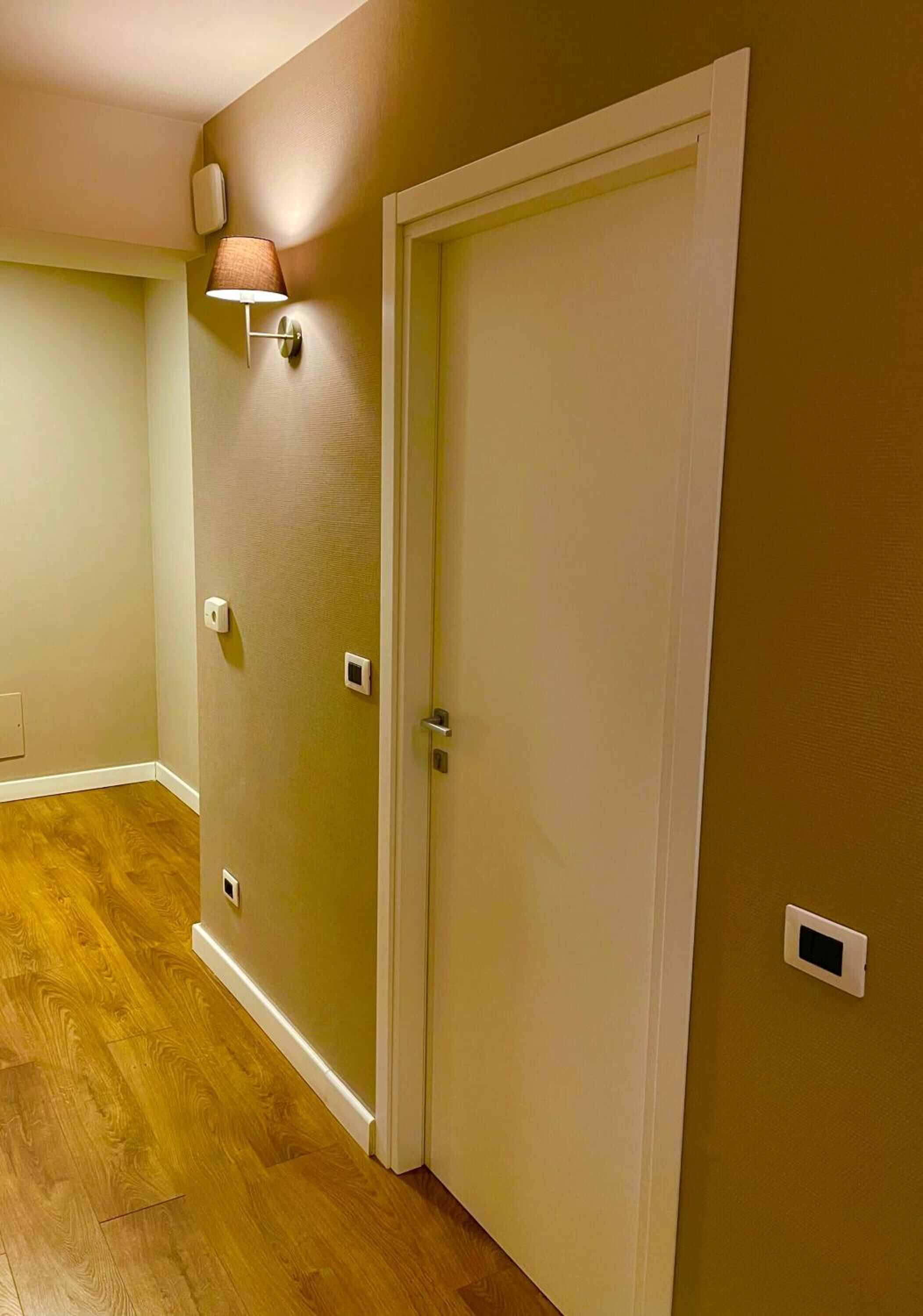 A hallway with a wooden floor, a closed door, and a wall-mounted light fixture.