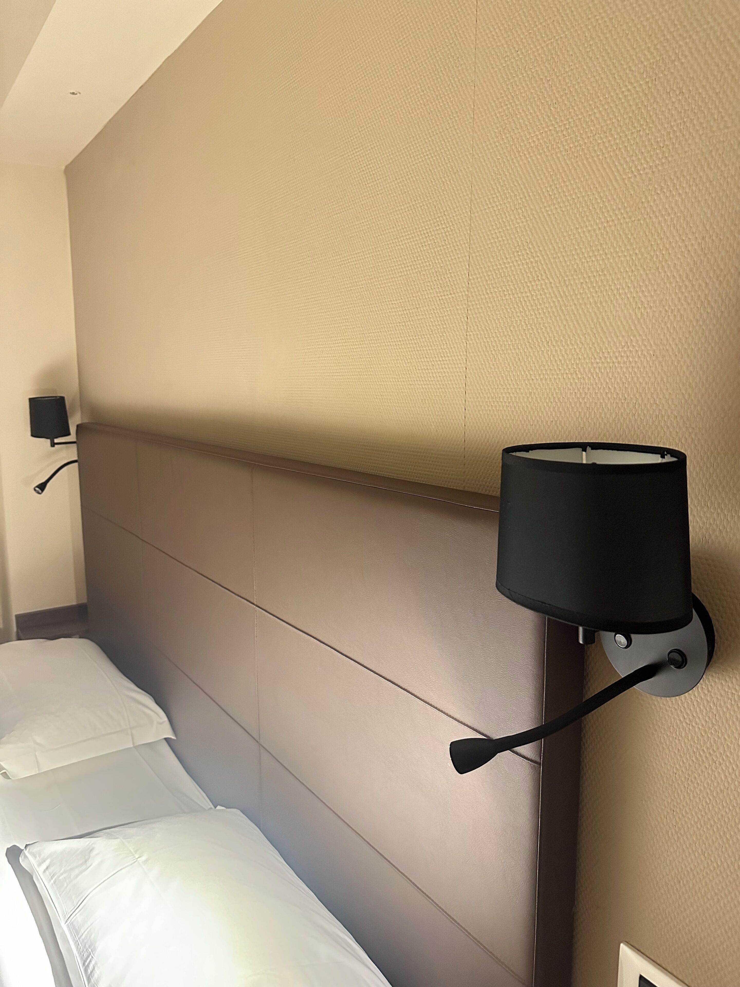 Une chambre d’hôtel avec une tête de lit, une lampe fixée au mur et un lit avec du linge de lit blanc.