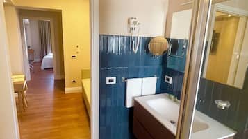 A modern bathroom with a shower, a sink, and a mirror. There is a door leading to a bedroom with a bed and a chair.