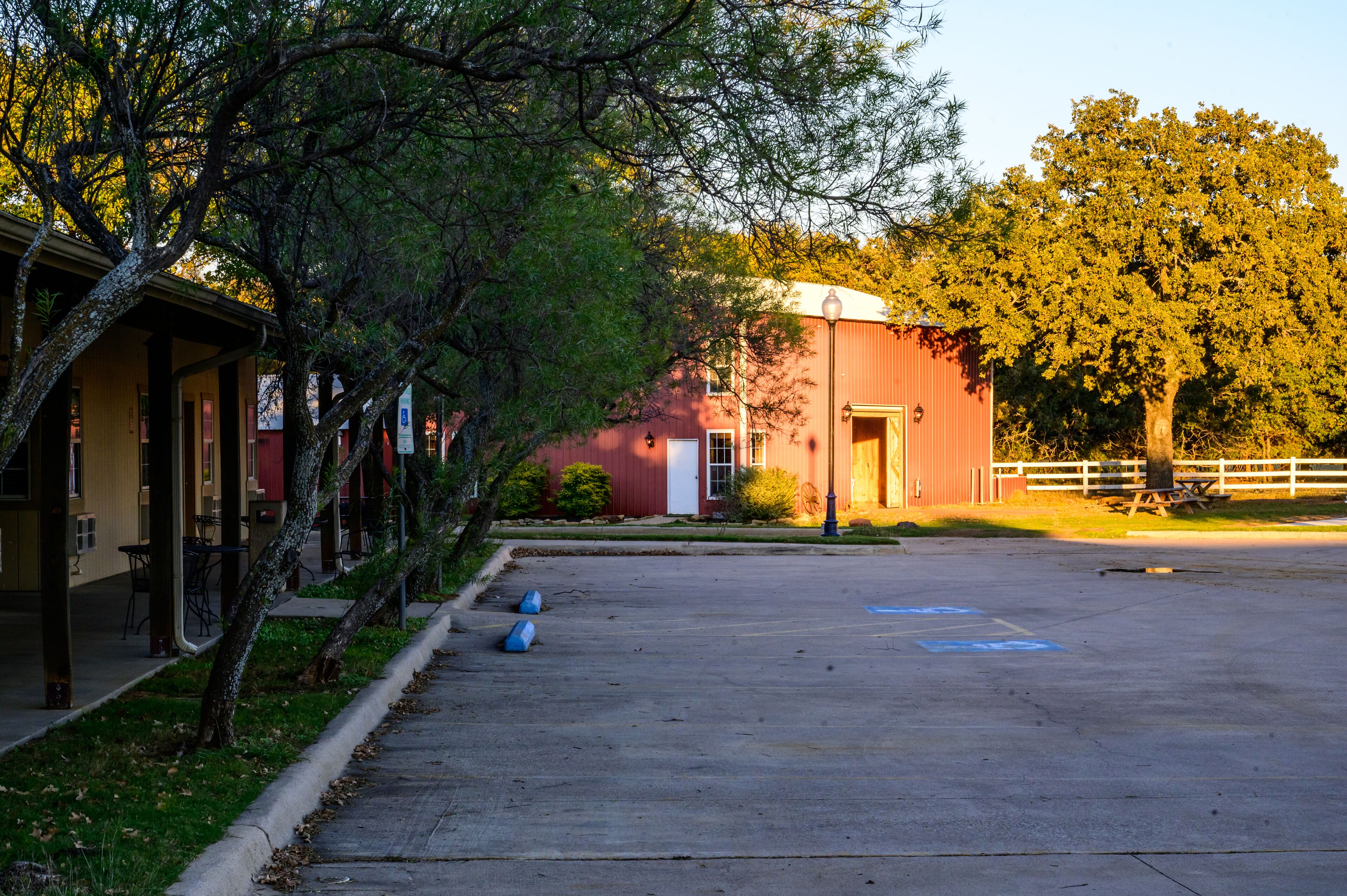A parking lot with a red building, trees, and a white fence.