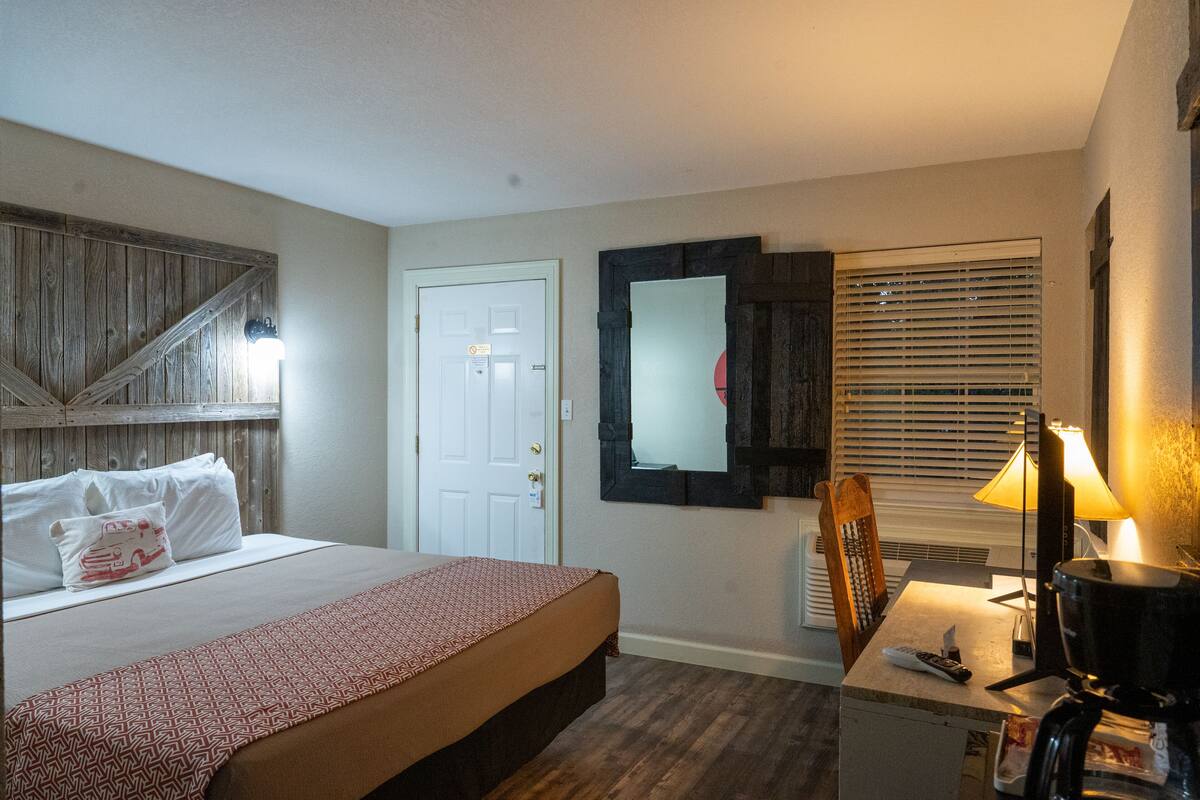 Ein Hotelzimmer mit Bett, Schreibtisch mit Lampe, Kaffeemaschine, Stuhl und einer Tür.