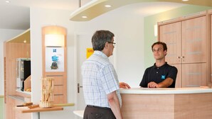 A reception area with a wooden reception desk, a counter, a wall-mounted lamp, and a poster. - Ibis Budget Augsburg Gersthofen (Gersthofen)