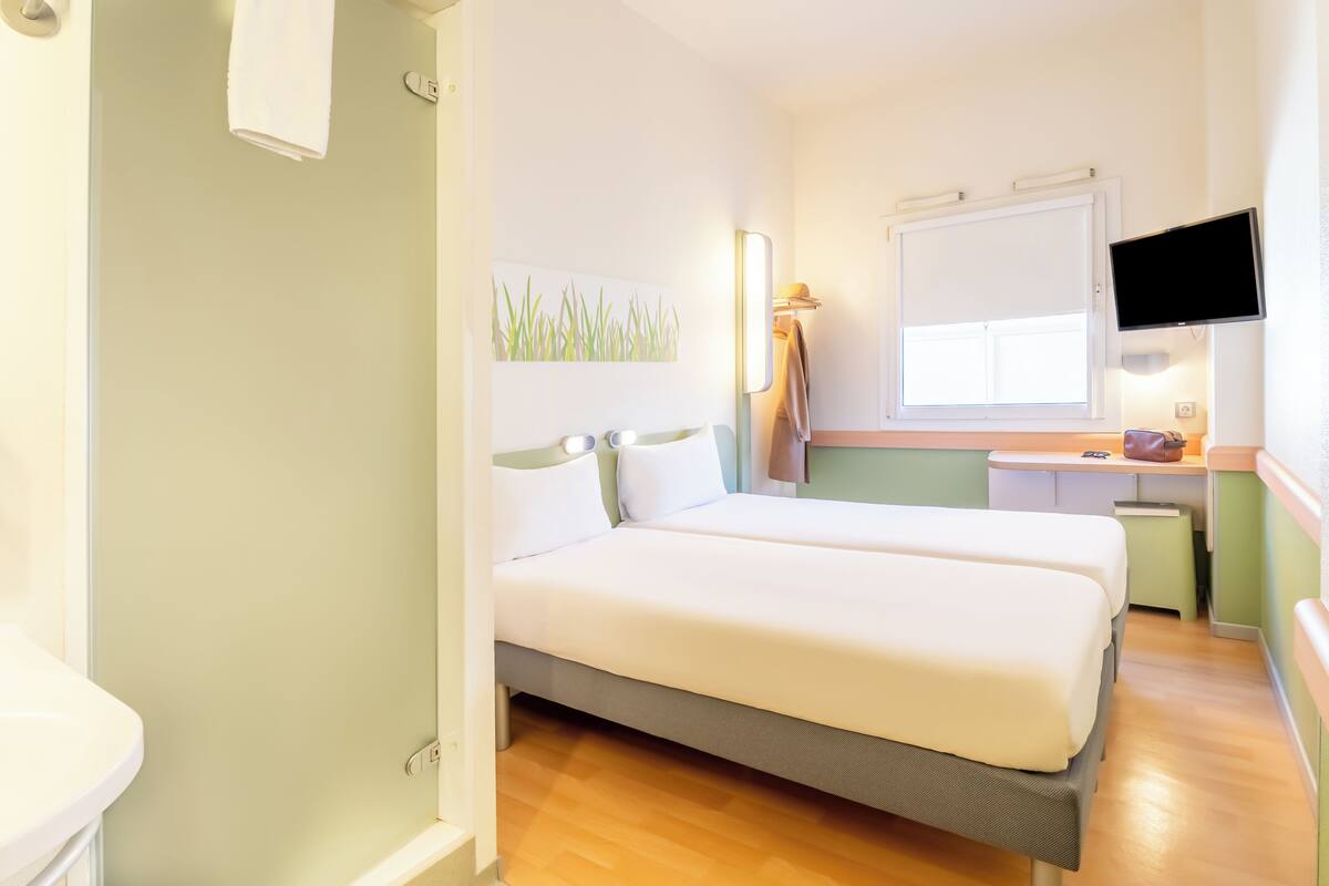A hotel room with two beds, a desk, a television, and a window.
