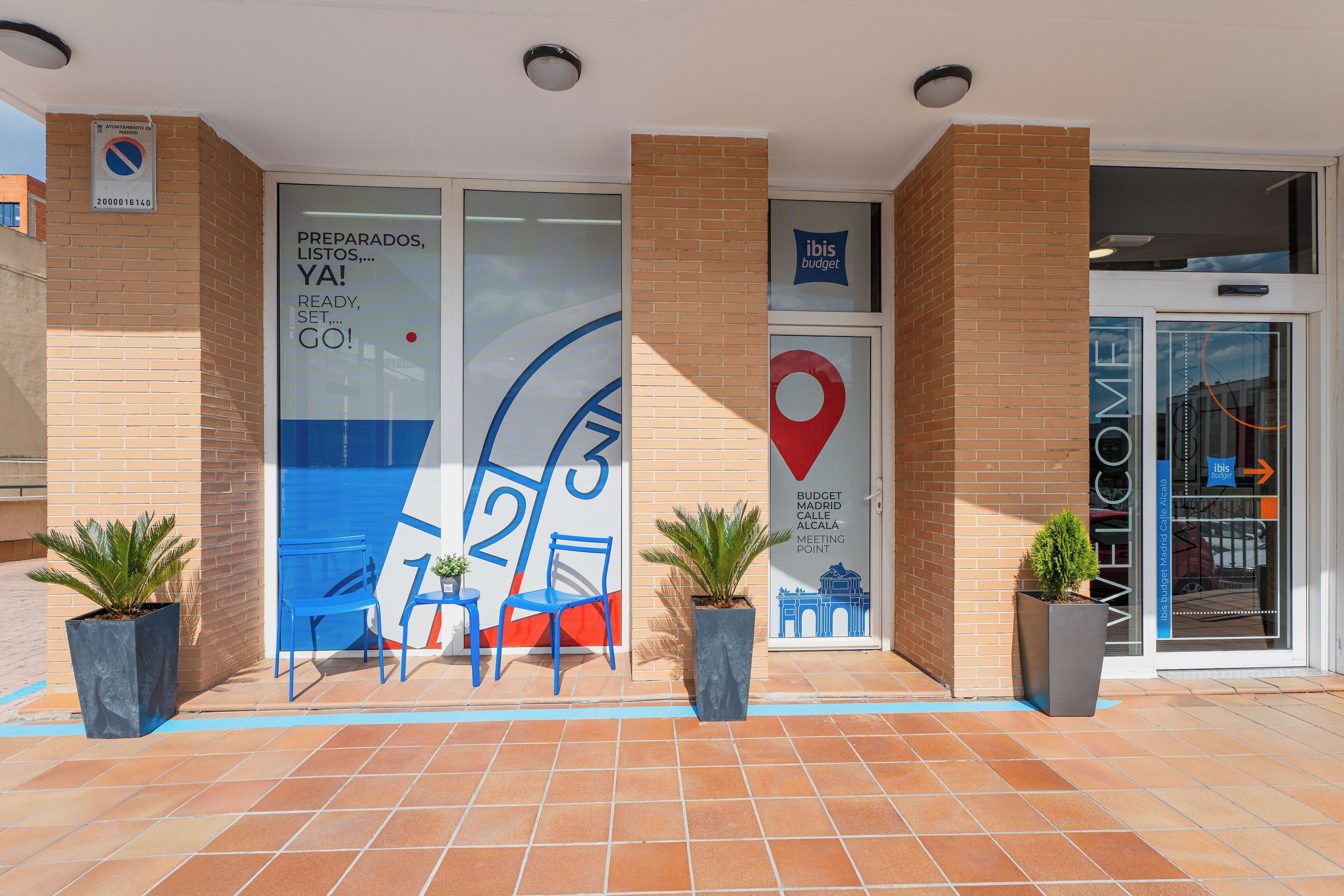 A hotel entrance with a glass door, blue chairs, potted plants, and a sign with a red location pin.