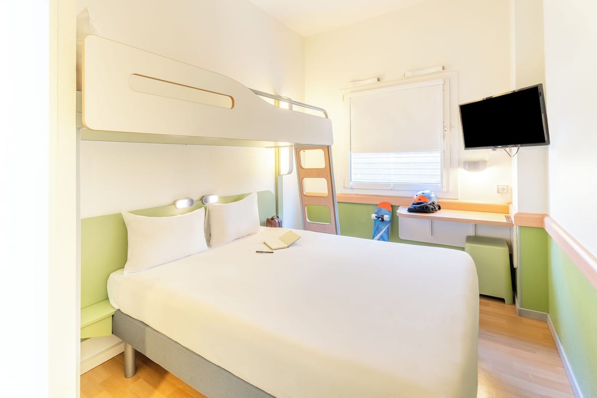 A bedroom with a bunk bed, a desk with a chair, a television mounted on the wall, and a window with blinds.