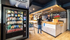 A modern reception area with a vending machine stocked with snacks and drinks, a wooden counter, and a chalkboard menu. - ibis budget Madrid Calle Alcalá (Madrid)