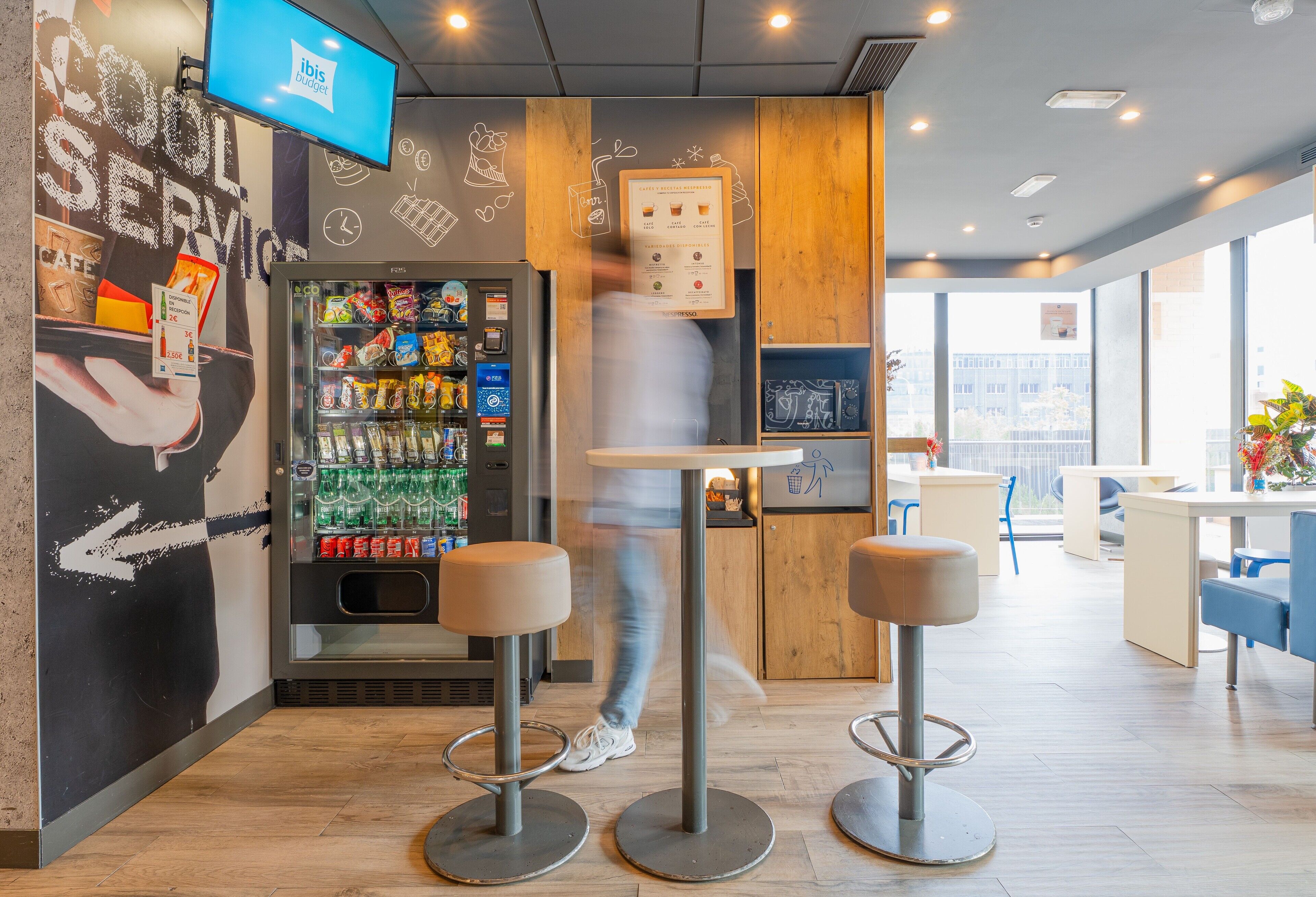 A modern bar area with a vending machine, stools, and a television screen displaying 'ibis budget'.