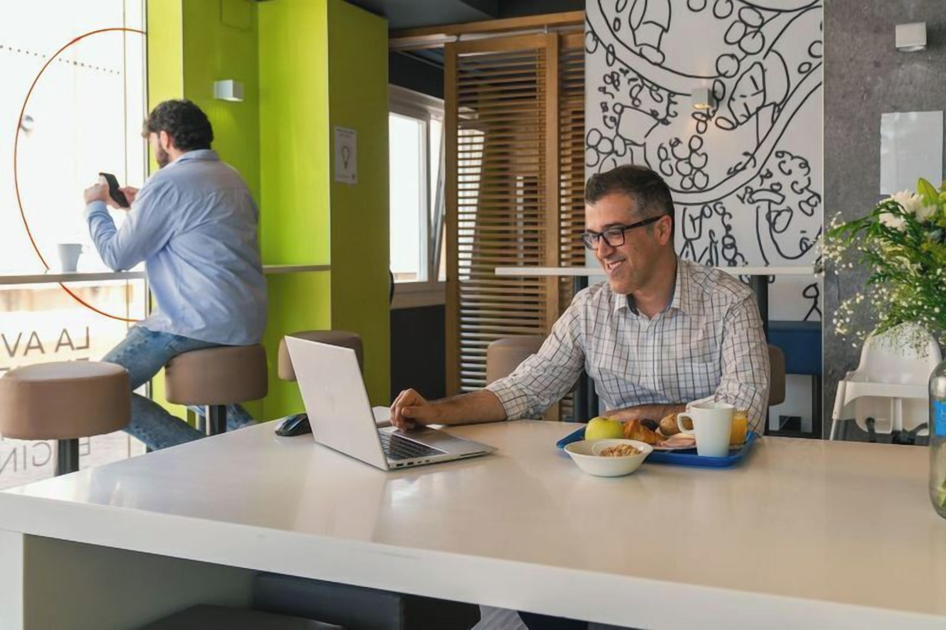 A modern office space with a person working on a laptop at a white table, another person in the background, and a wall with a black and white abstract design.