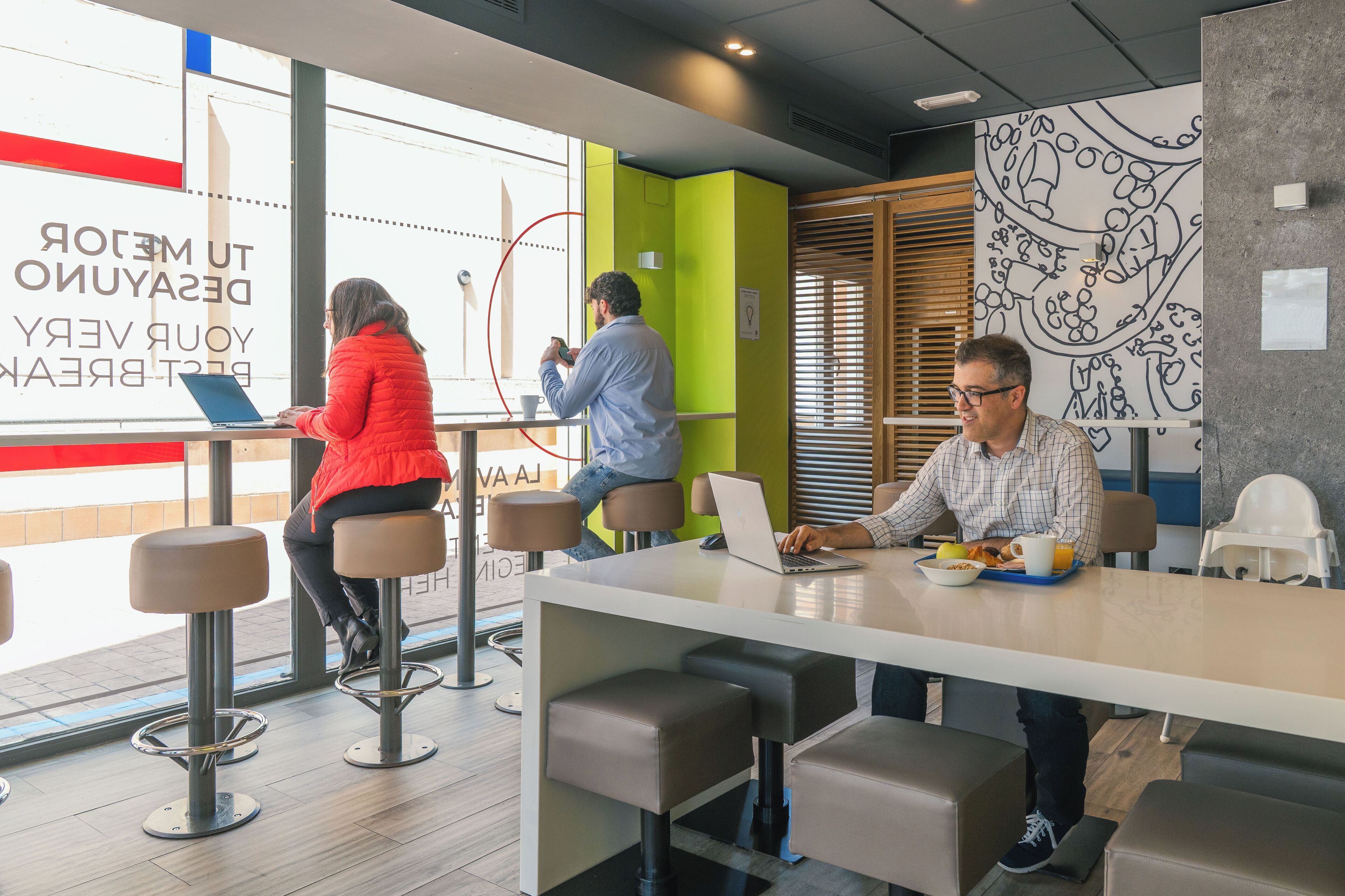 Un espace de bureau moderne doté de plusieurs postes de travail, d’une table commune où l’on prend le petit-déjeuner, et d’une grande fenêtre laissant entrer la lumière naturelle.