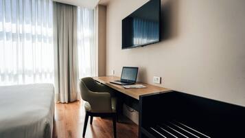 Habitación de hotel con una cama, un escritorio con una computadora portátil, una silla, un televisor montado en la pared y una ventana con cortinas.