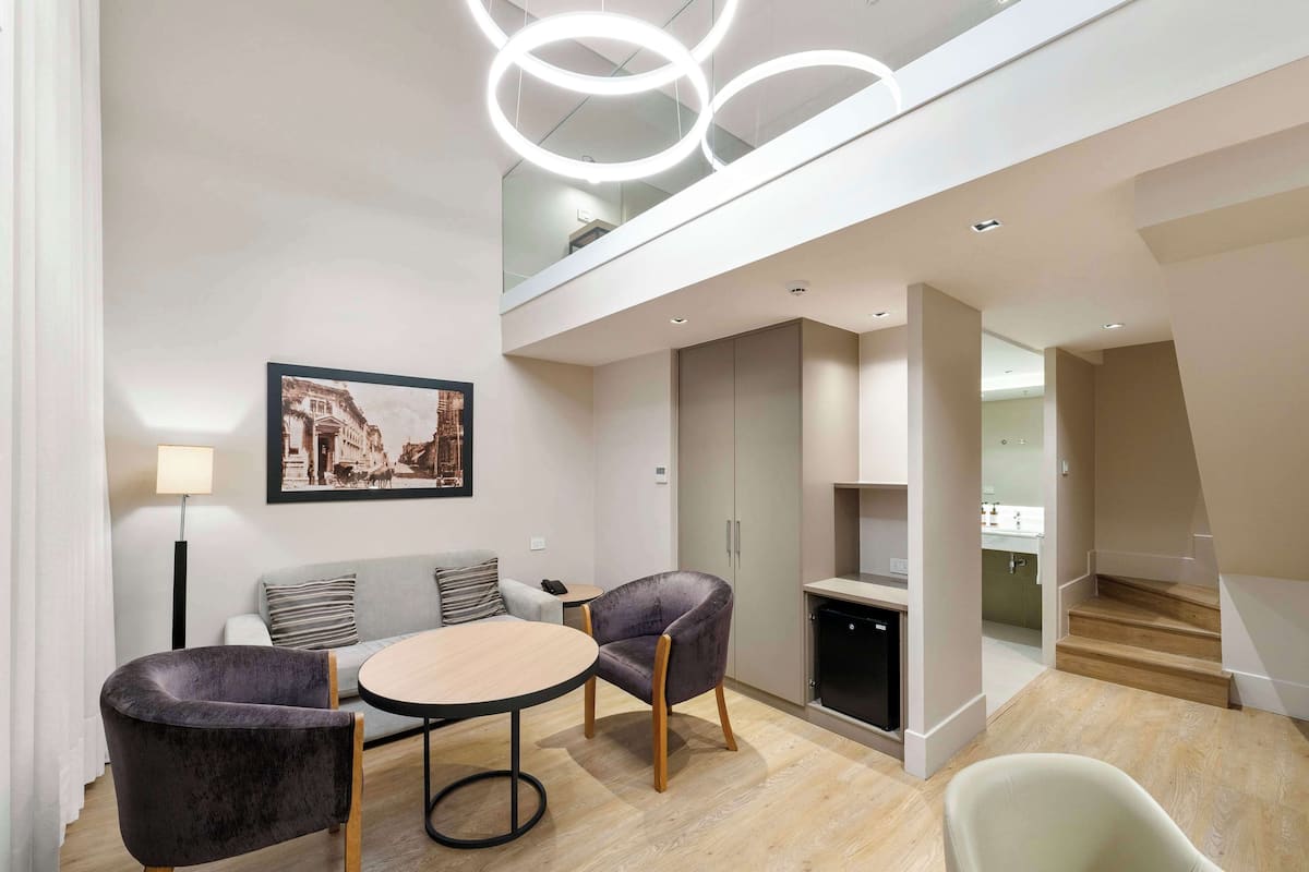 A modern living room with a round table, two armchairs, a sofa, and a framed cityscape picture. There is a staircase leading to a kitchen area.