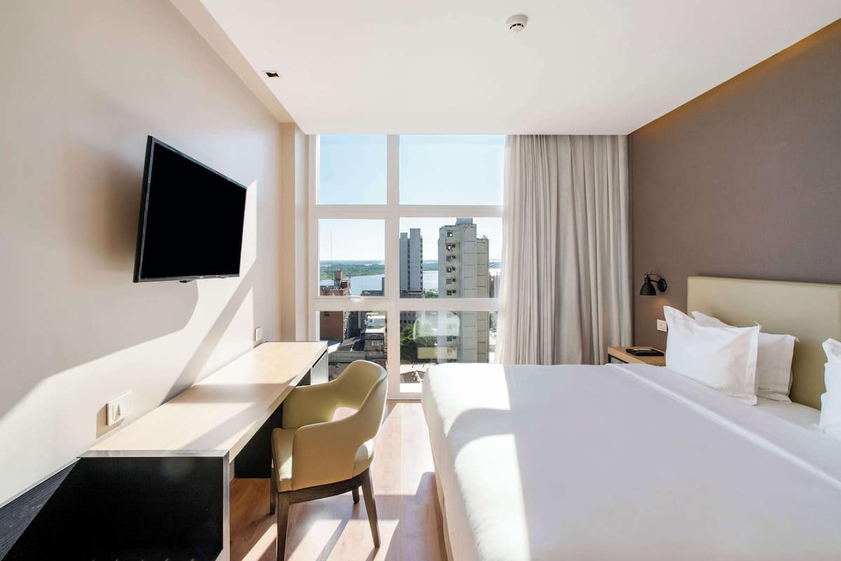 A hotel room with a bed, a desk, a chair, a TV, and a large window offering a city view.