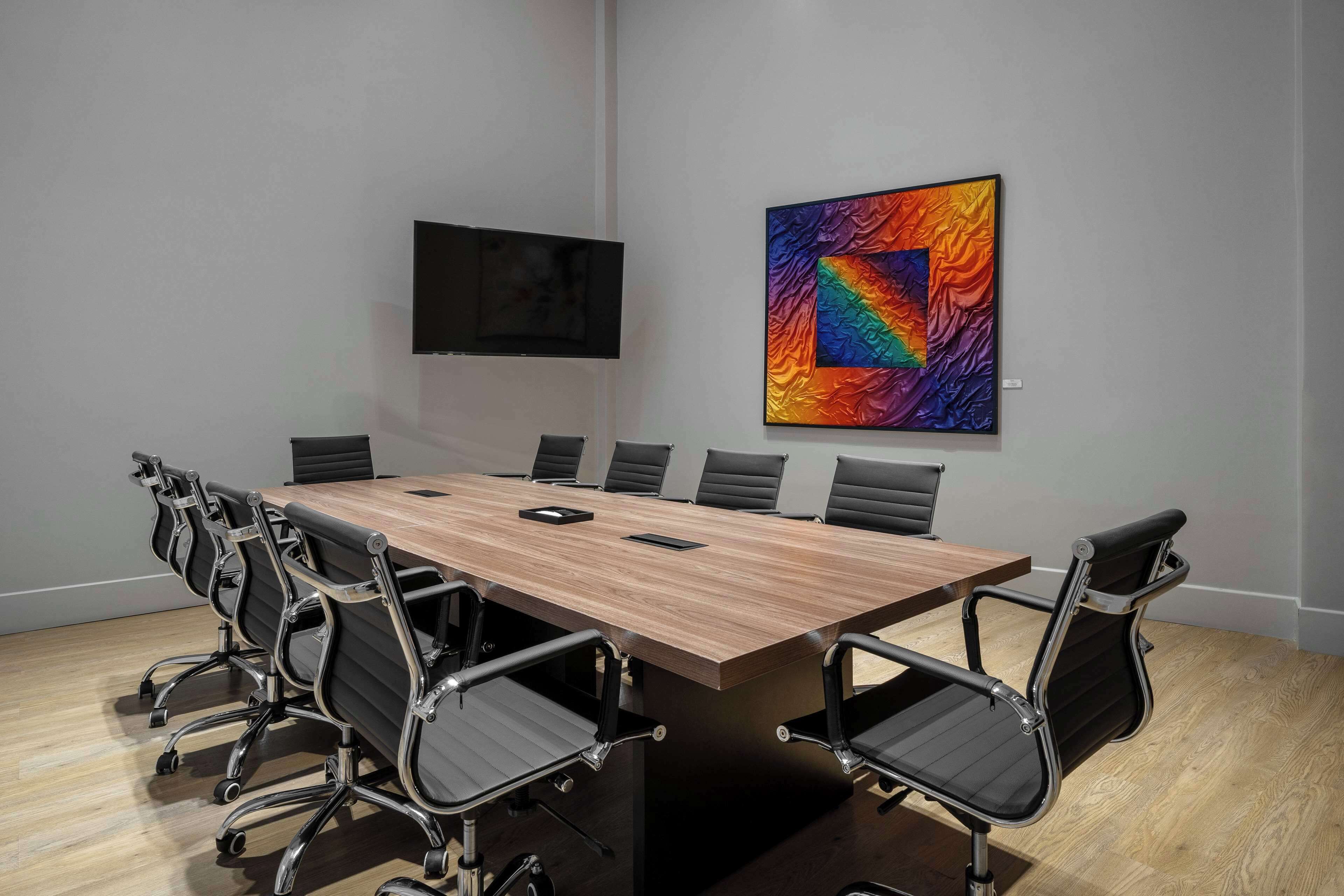 Un salón de conferencias con una larga mesa de madera, sillas negras, un televisor de pantalla plana y un cuadro abstracto de colores en la pared.