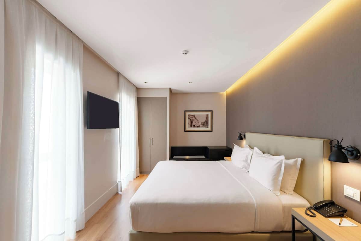 A modern hotel room with a large bed, a bedside table with a lamp, a flat-screen TV mounted on the wall, and a framed picture above the bed.