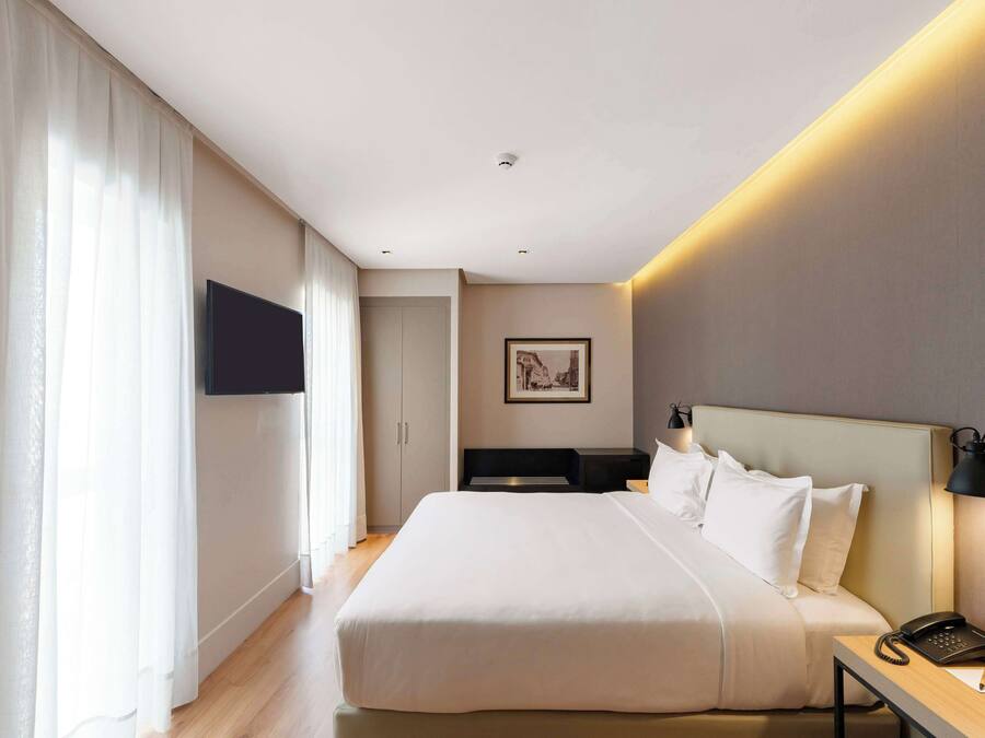 A modern hotel room with a large bed, a bedside table with a lamp, a flat-screen TV mounted on the wall, and a framed picture above the bed.