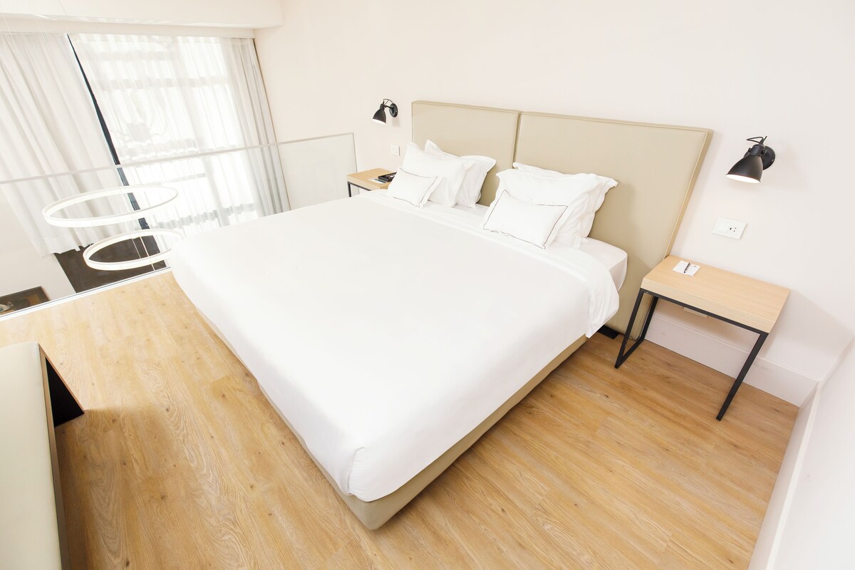 A modern hotel room with a large bed, wooden flooring, a desk, and a nightstand.