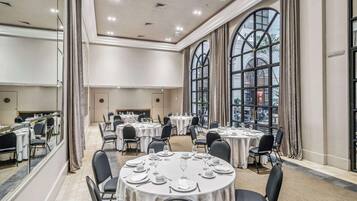 A spacious banquet hall with round tables set for dining, large windows with curtains, and a high ceiling with recessed lighting.
