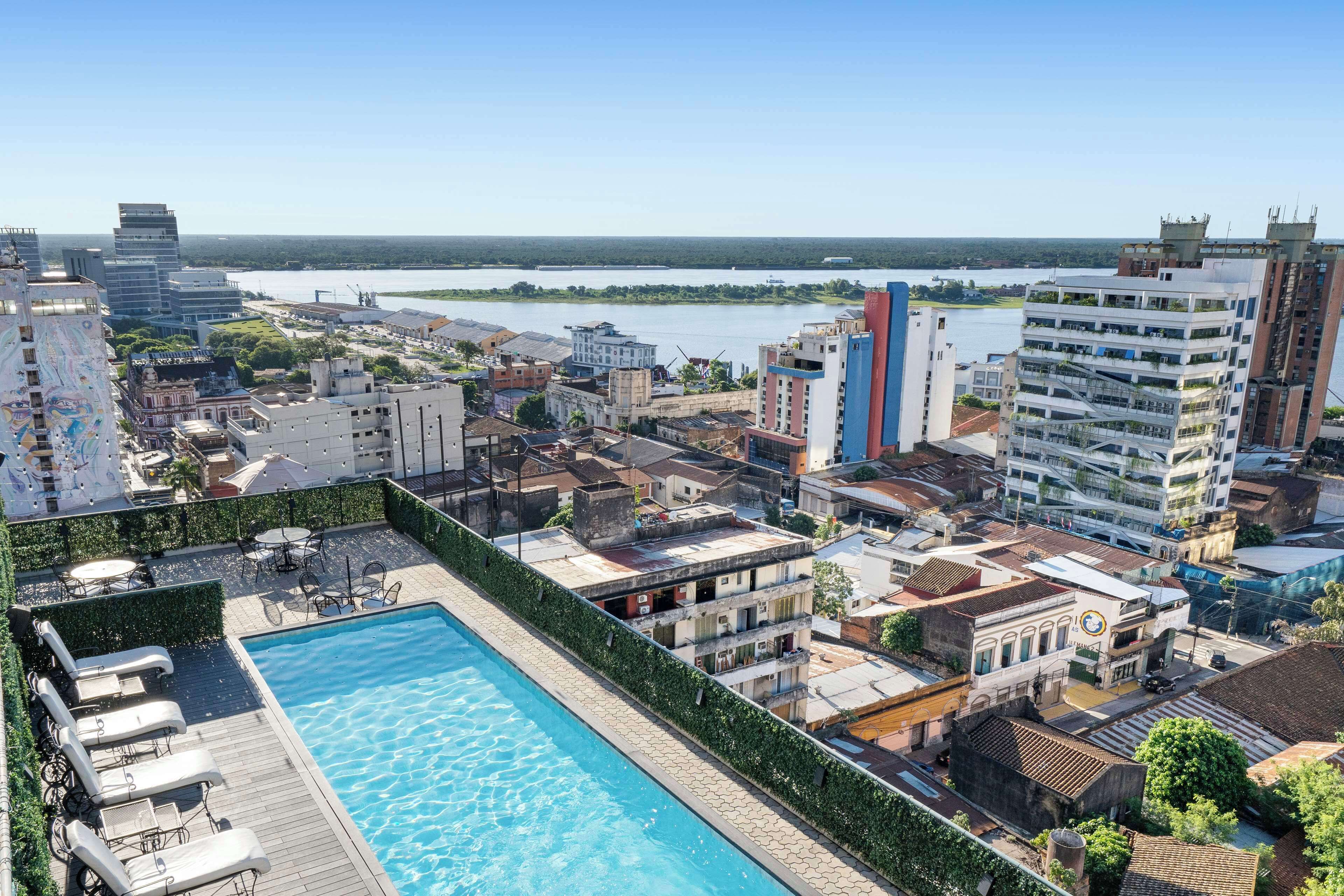 A rooftop pool with lounge chairs, surrounded by urban buildings and a river in the background.