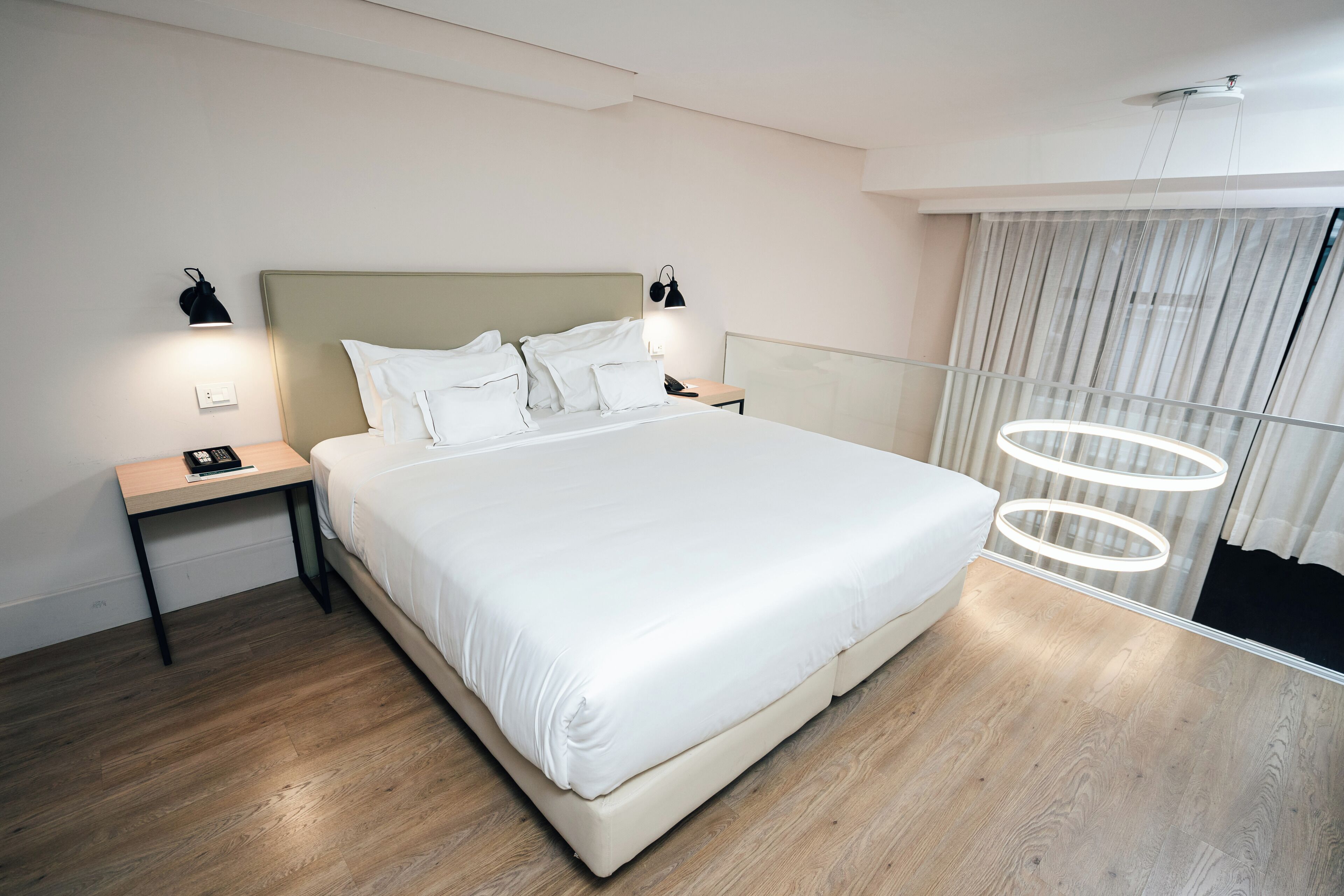 A modern hotel room with a large bed, bedside tables, and a view of the city through large windows.