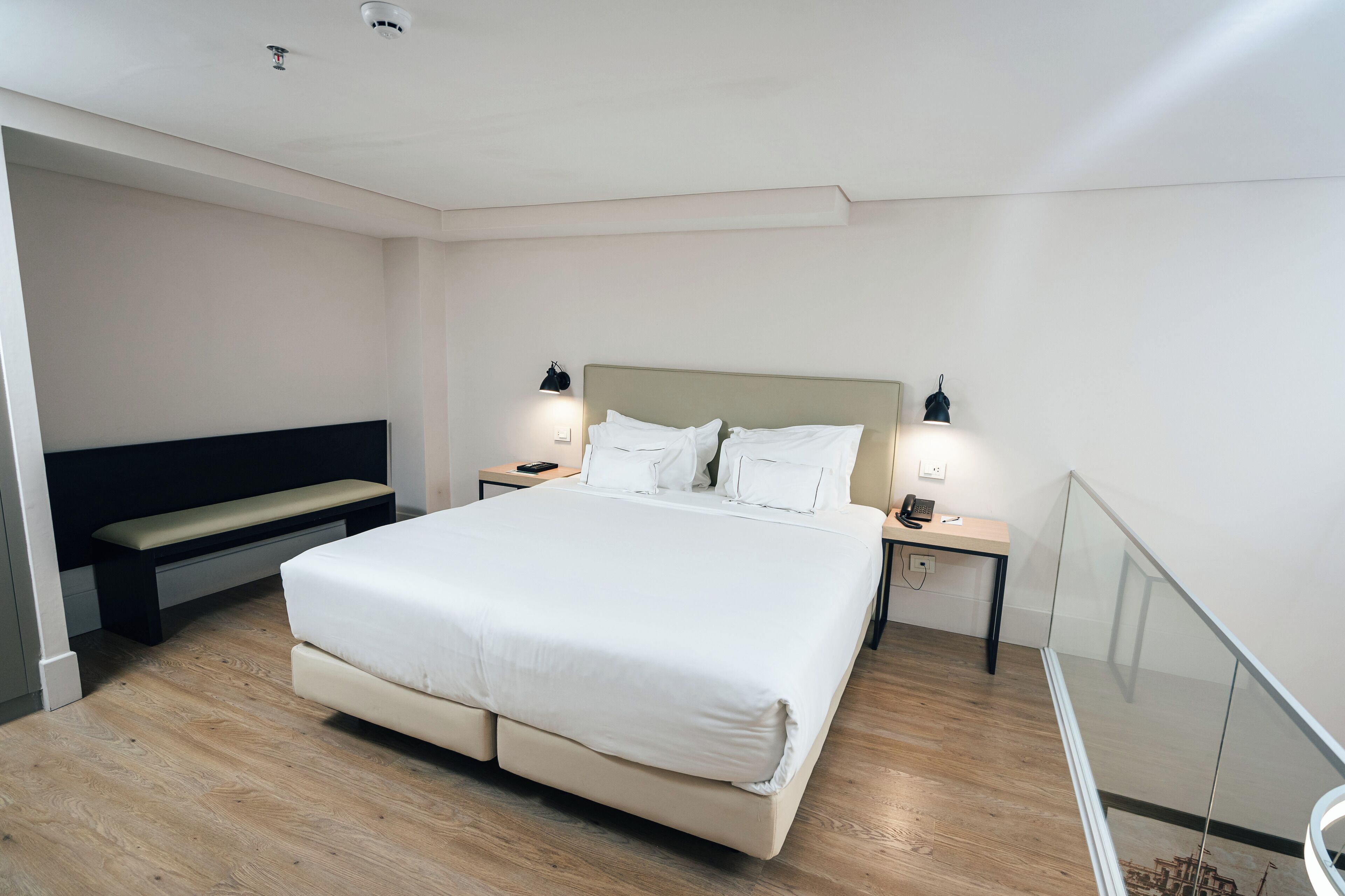 Una habitación de hotel moderna con una cama grande, mesitas de noche, un banco y una barandilla de vidrio.