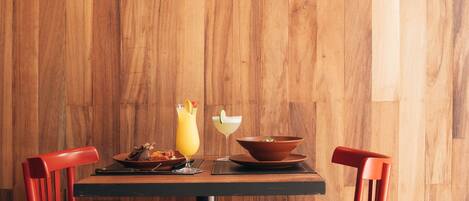 A wooden wall with the name "LA MARIA" and "COCINA Y CARBON" written on it. There is a table with two red chairs and a yellow drink on it.
