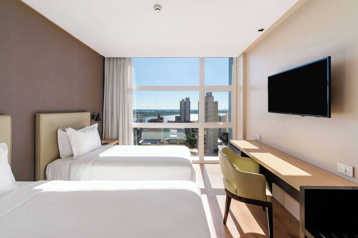 A hotel room with two beds, a desk, a chair, a TV, and a large window offering a city view.
