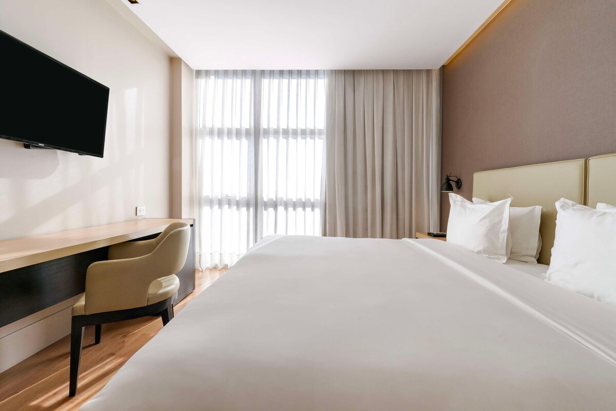 Hotellihuone, jossa on suuri sänky, työpöytä, tuoli, televisio ja ikkuna, jossa on verhot.