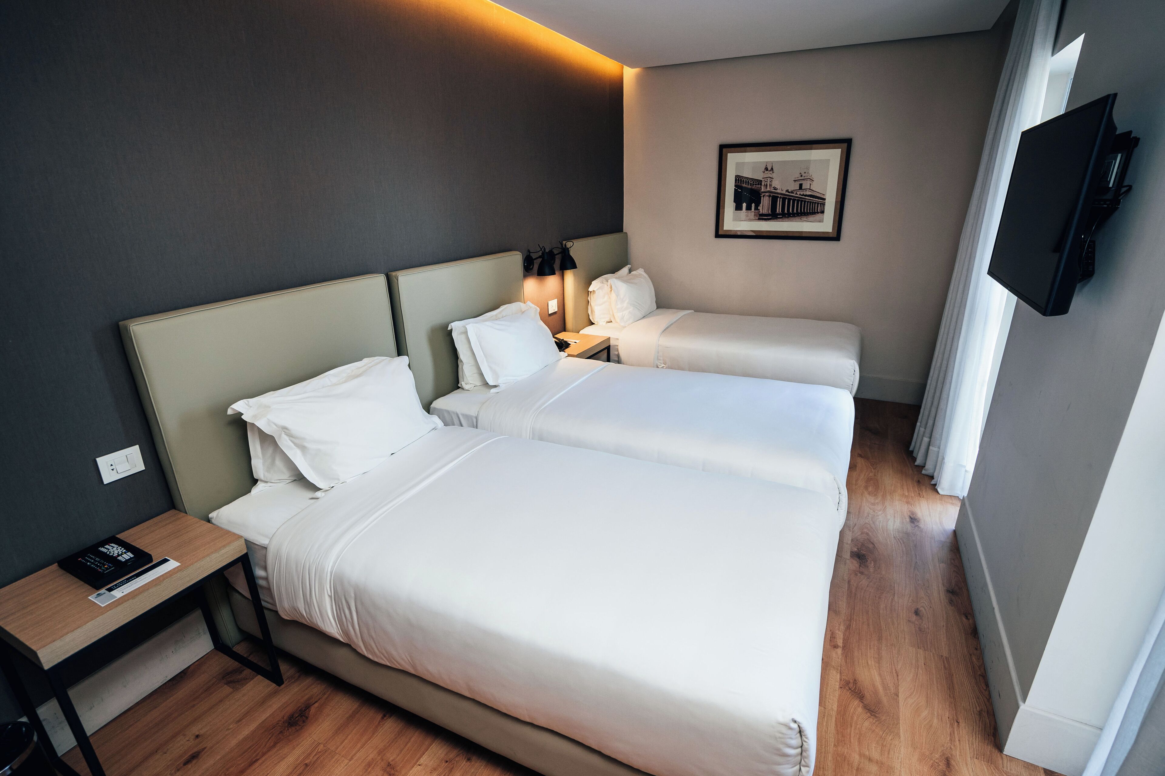 A hotel room with two beds, a bedside table with a remote control, a framed picture on the wall, and a TV mounted on the wall.
