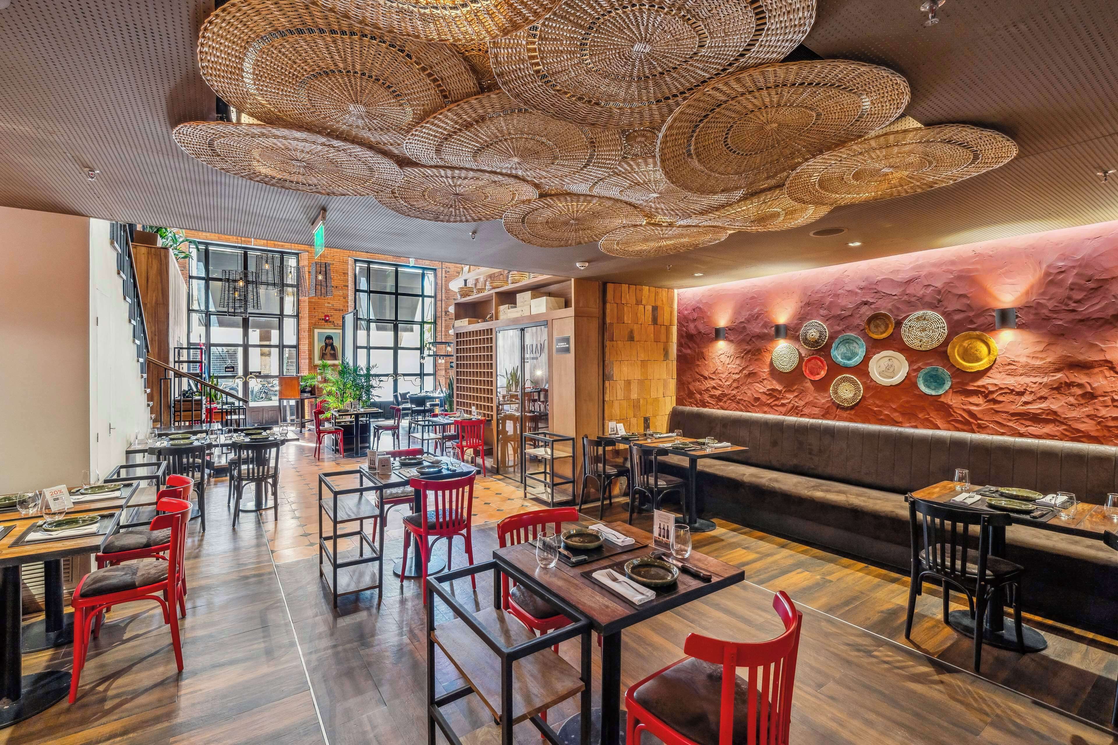Un restaurante con mesas de madera y sillas rojas, una pared de ladrillo adornada con platos decorativos y lámparas de techo tejidas.