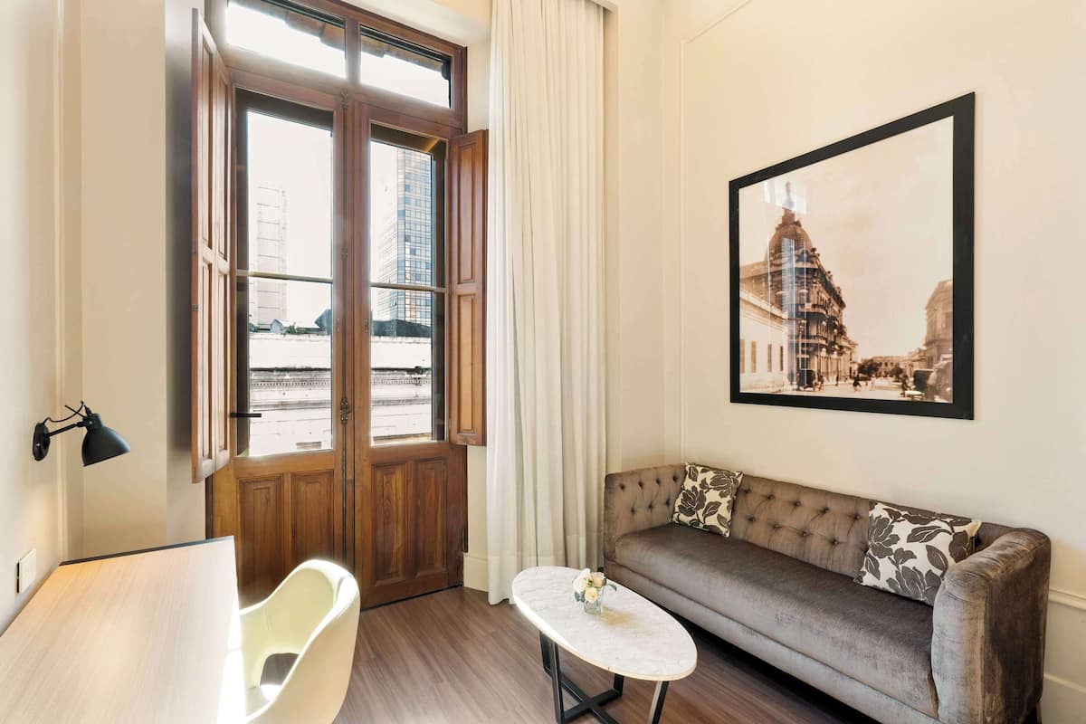 A room with a wooden door, a white chair, a sofa with patterned cushions, a small table with a vase, and a framed picture on the wall.