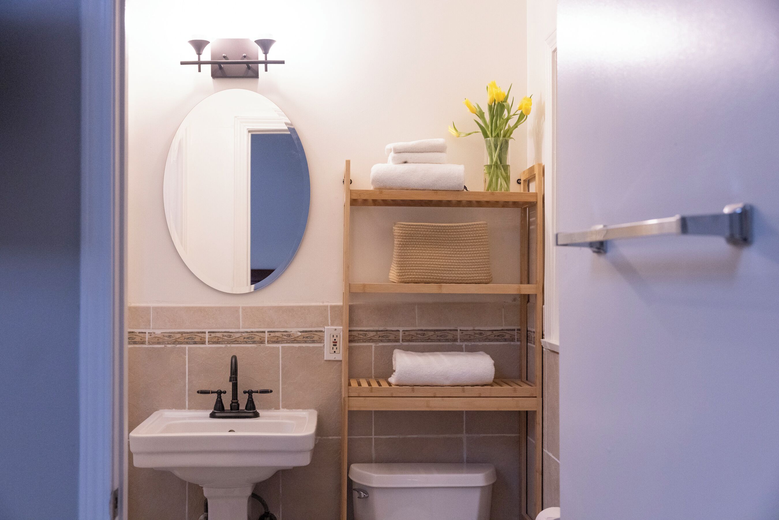 A bathroom with a sink, toilet, mirror, and shelves with towels and a vase of flowers.