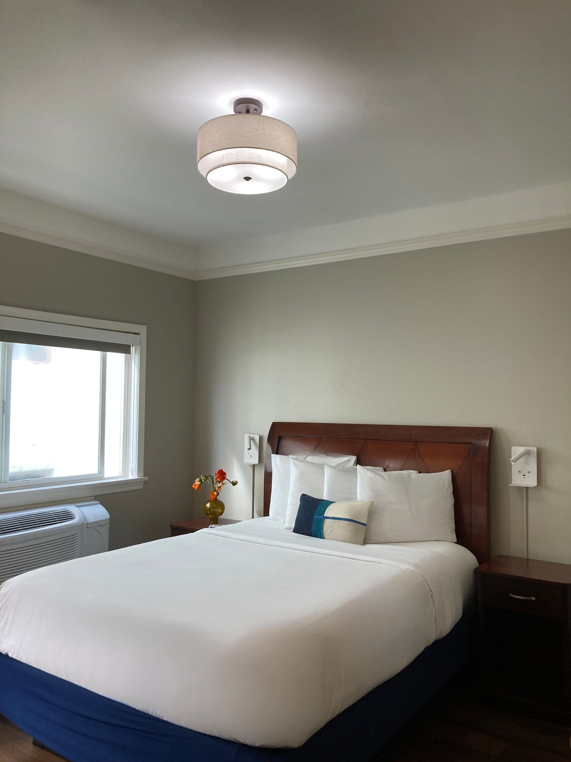 Una habitación de hotel con cama, mesita de noche, enchufe en la pared, ventana y lámpara en el techo.