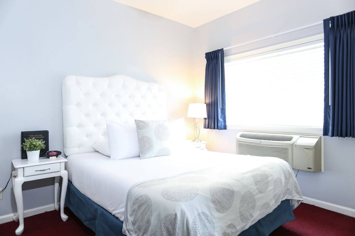 A hotel room with a tufted headboard, a bed with patterned bedding, a bedside table with a clock and a plant, a lamp, and a window with curtains.