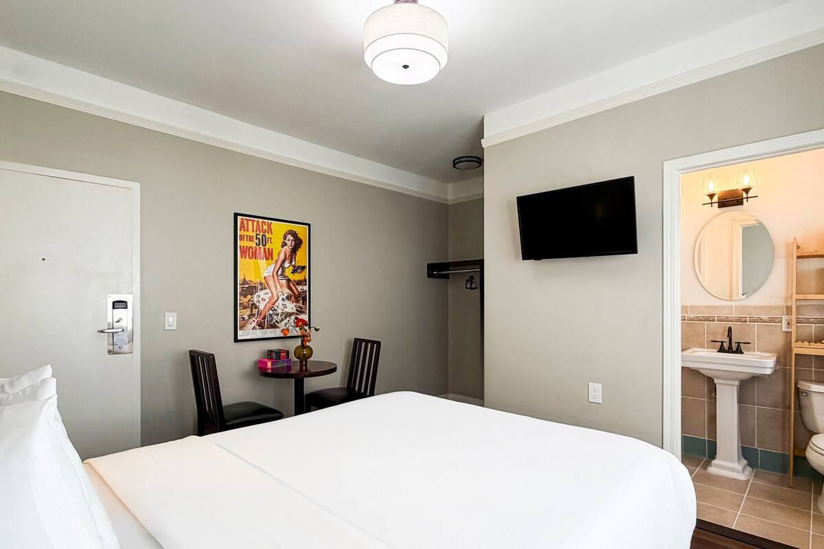 A hotel room with a bed, a television mounted on the wall, a poster on the wall, a small table with chairs, and a bathroom visible through an open door.