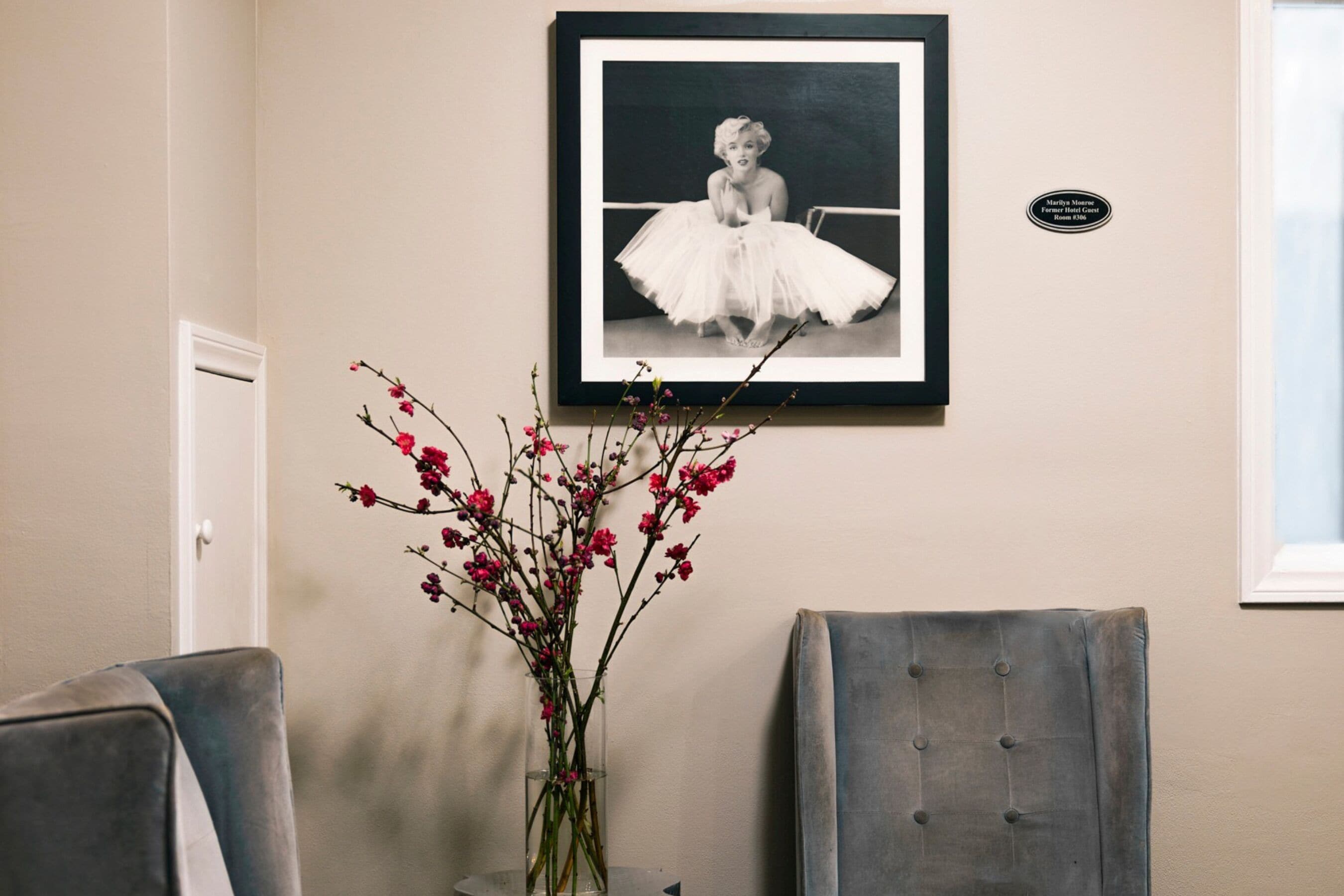 A framed photograph on the wall, a vase with pink flowers, and two chairs.