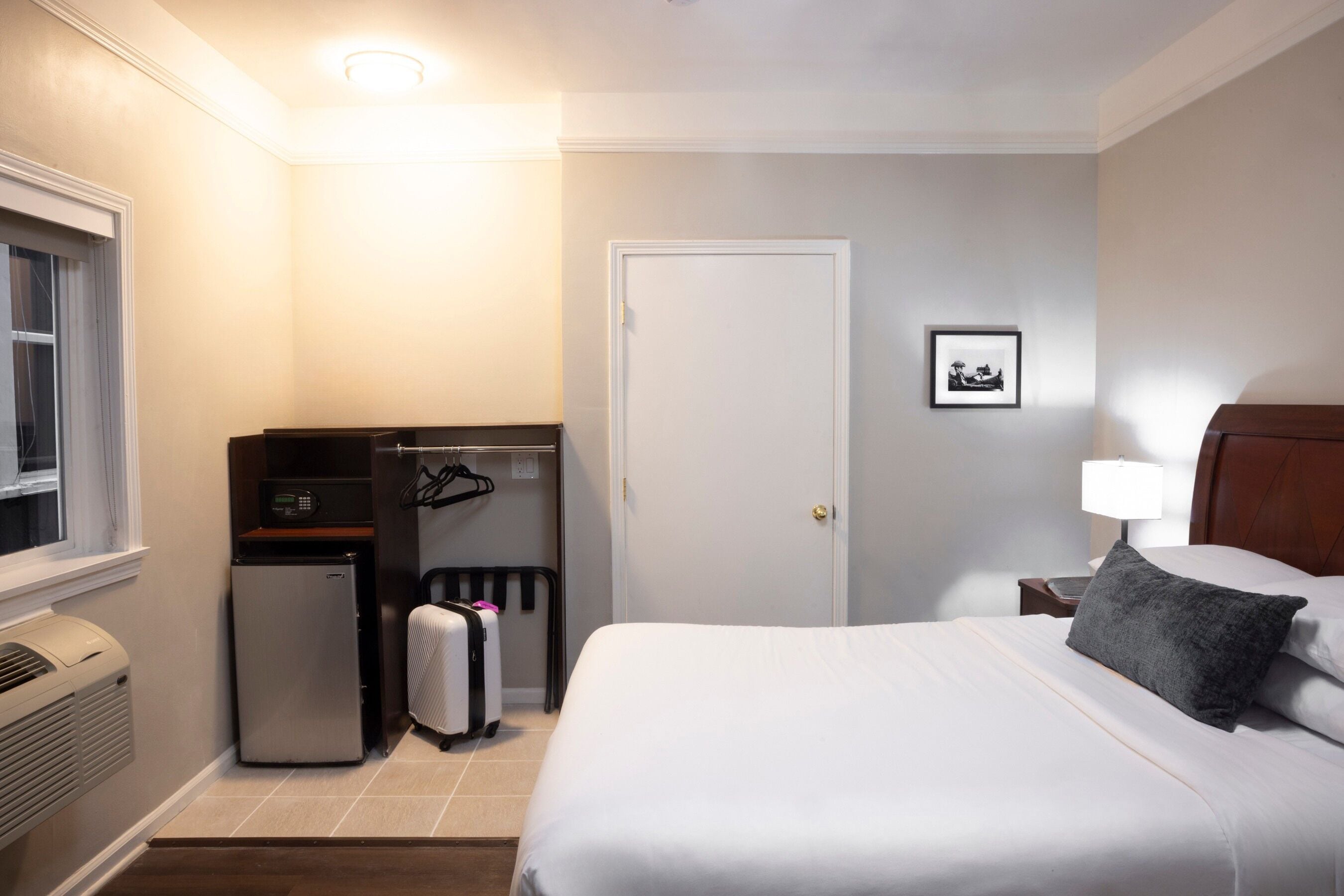Une chambre d’hôtel comprenant un lit, un petit réfrigérateur, un radiateur, une valise, un porte-manteau, une porte et une table de chevet avec une lampe.