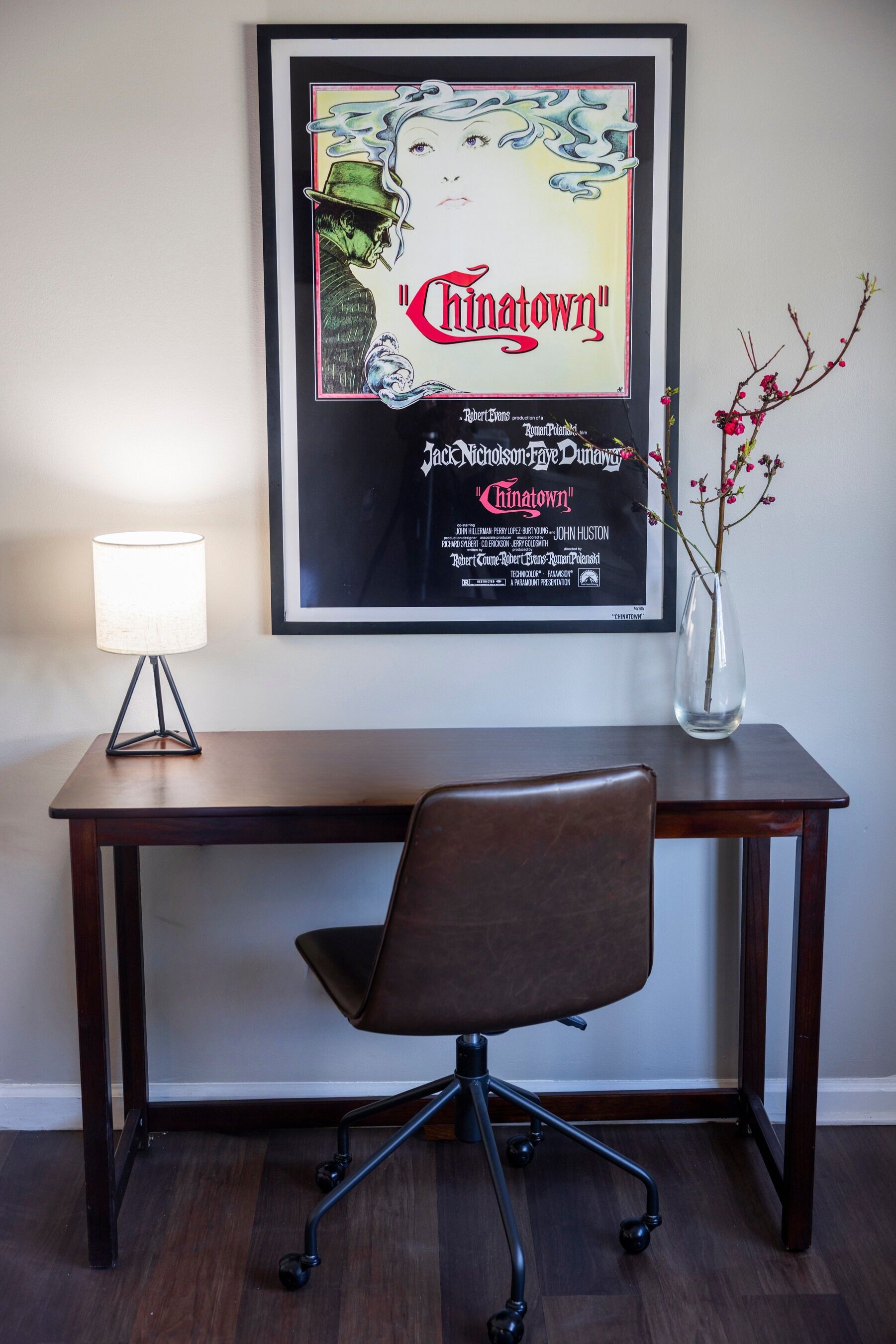 A framed movie poster of 'Chinatown' on a wall, a desk with a lamp, a chair, and a vase with branches.