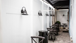 A narrow corridor with white panel walls, black wall-mounted lights, and a row of dark wooden chairs and tables. - The Hotel Hollywood (Los Angeles)