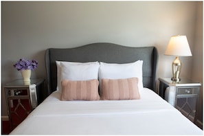 Cama bem arrumada com duas almofadas, abajur e um vaso com flores em uma mesa de cabeceira.