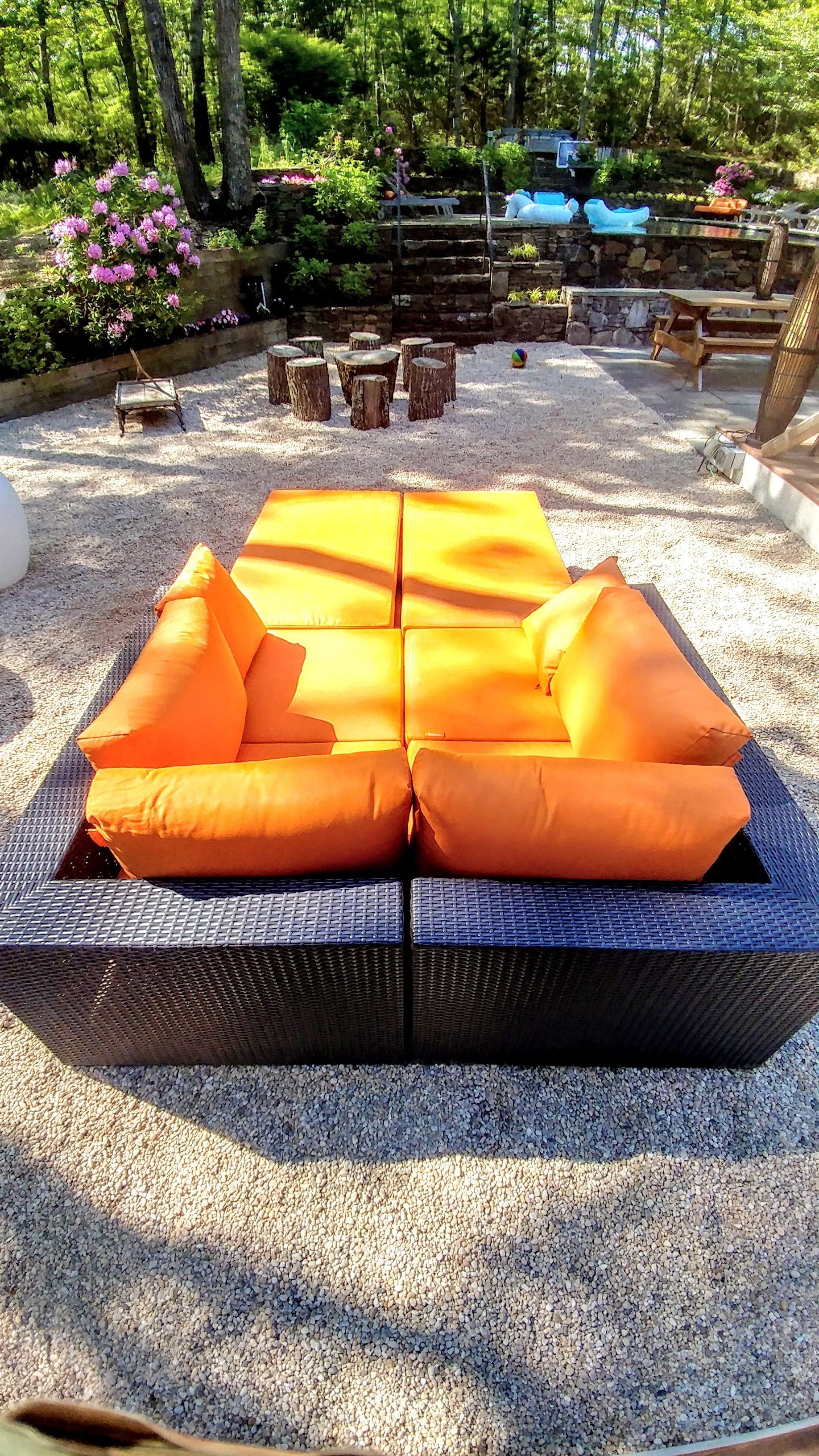 A wicker sofa with orange cushions in an outdoor setting with a gravel ground, surrounded by trees and a stone wall.