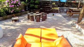 A wicker sofa with orange cushions in an outdoor setting with a gravel ground, surrounded by trees and a stone wall.