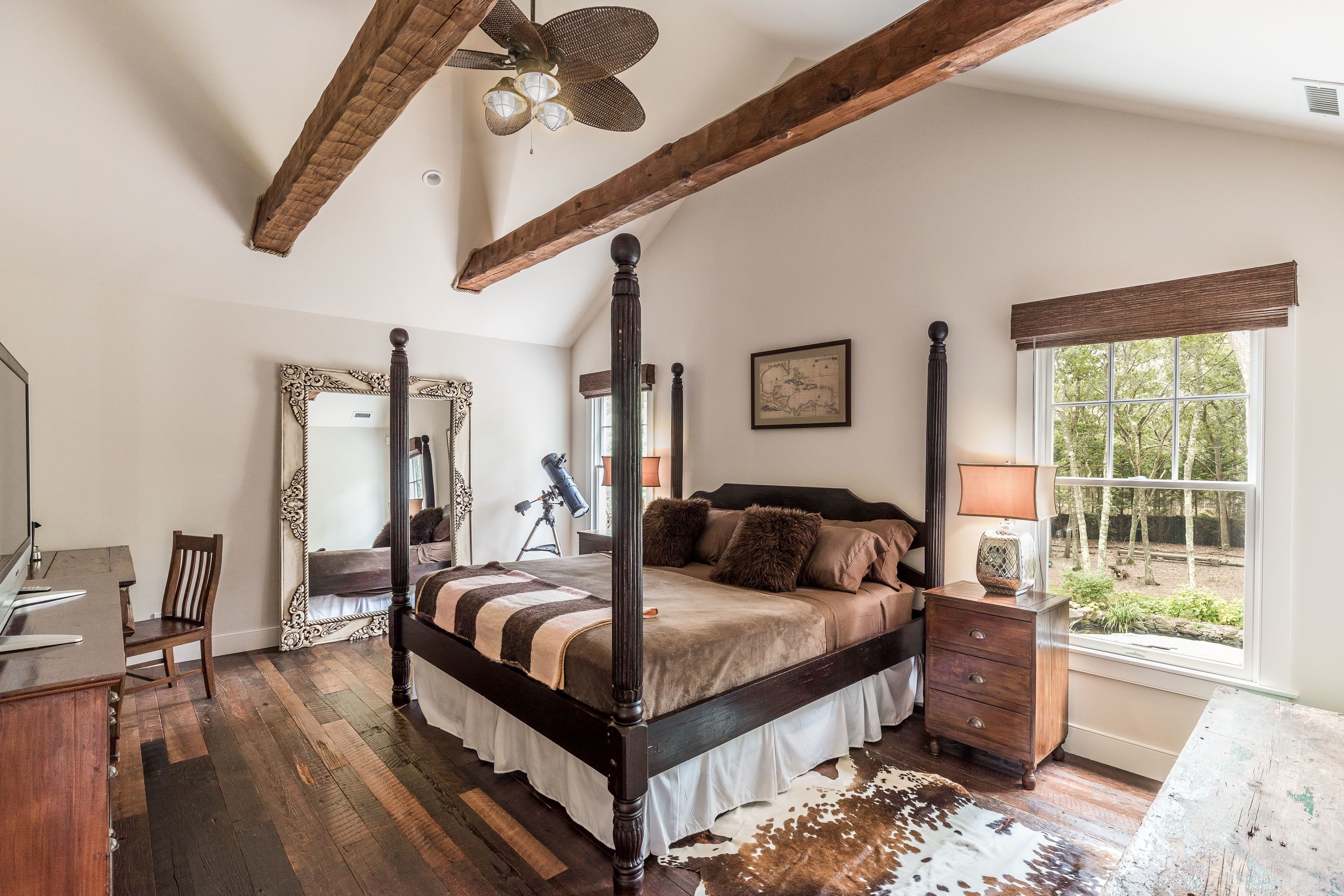 Un lit à baldaquin au revêtement foncé, un bureau en bois avec une chaise, un grand miroir et une fenêtre donnant sur les arbres.