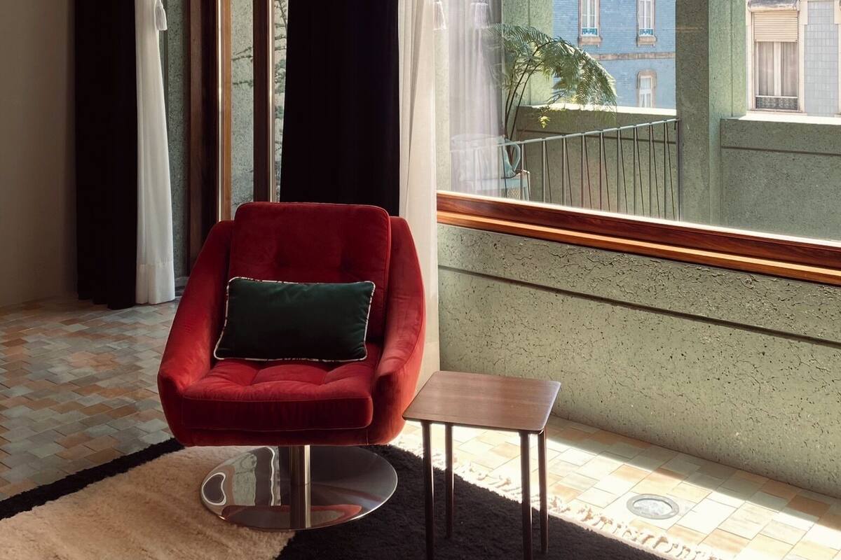 Una habitación con un sillón rojo, una mesita y vistas a edificios a través de un ventanal con cortinas blanco y negro.