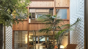 Un edificio moderno con fachada de madera, ventanas amplias y un patio con plantas en macetas y zonas de estar.