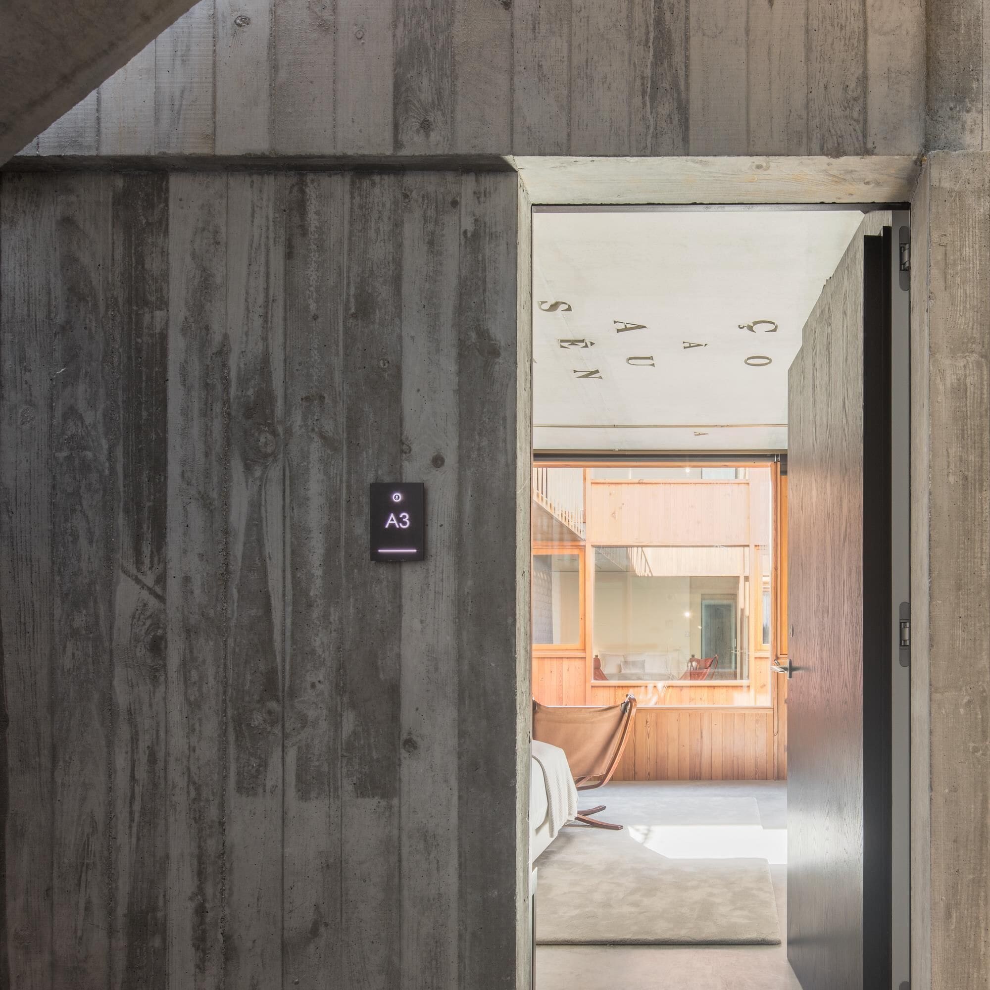 Un interior moderno con una pared de hormigón y una puerta que da a un cuarto con suelo de madera y una cama.