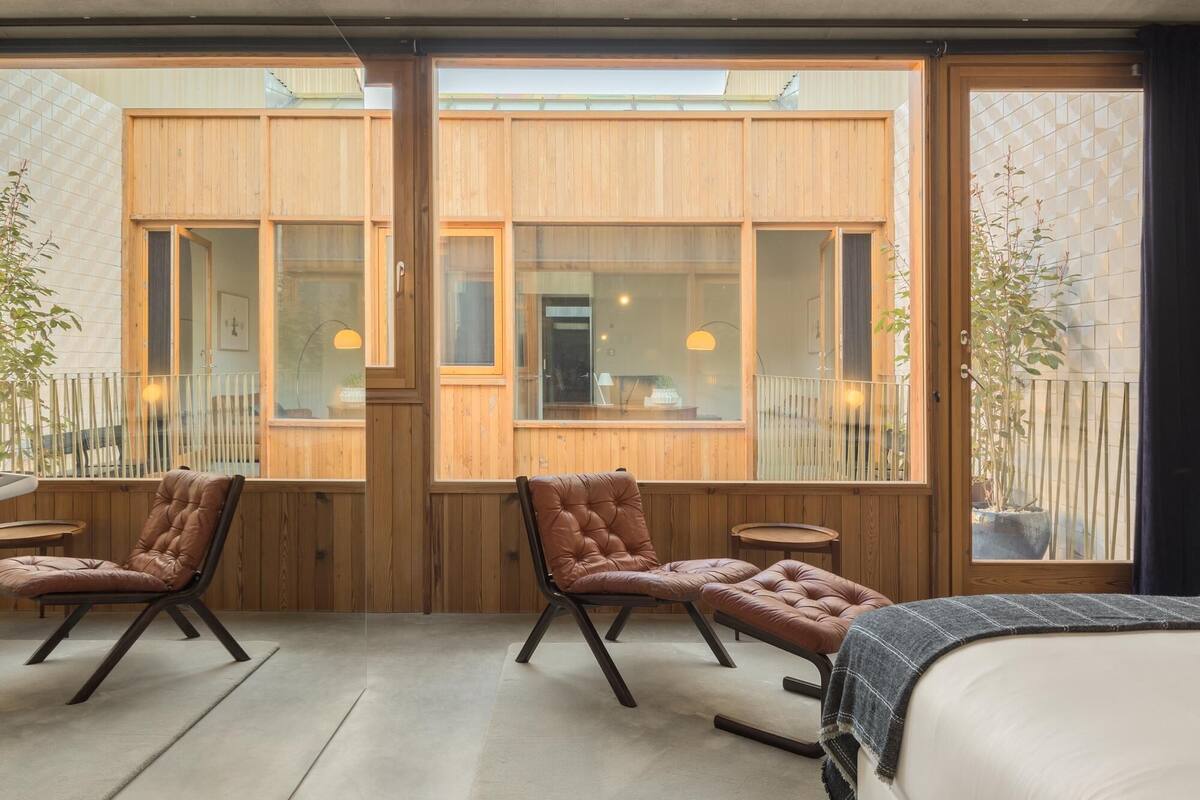 Una habitación moderna con una cama grande, dos sillones de cuero, una mesita y ventanas amplias con vistas al exterior.