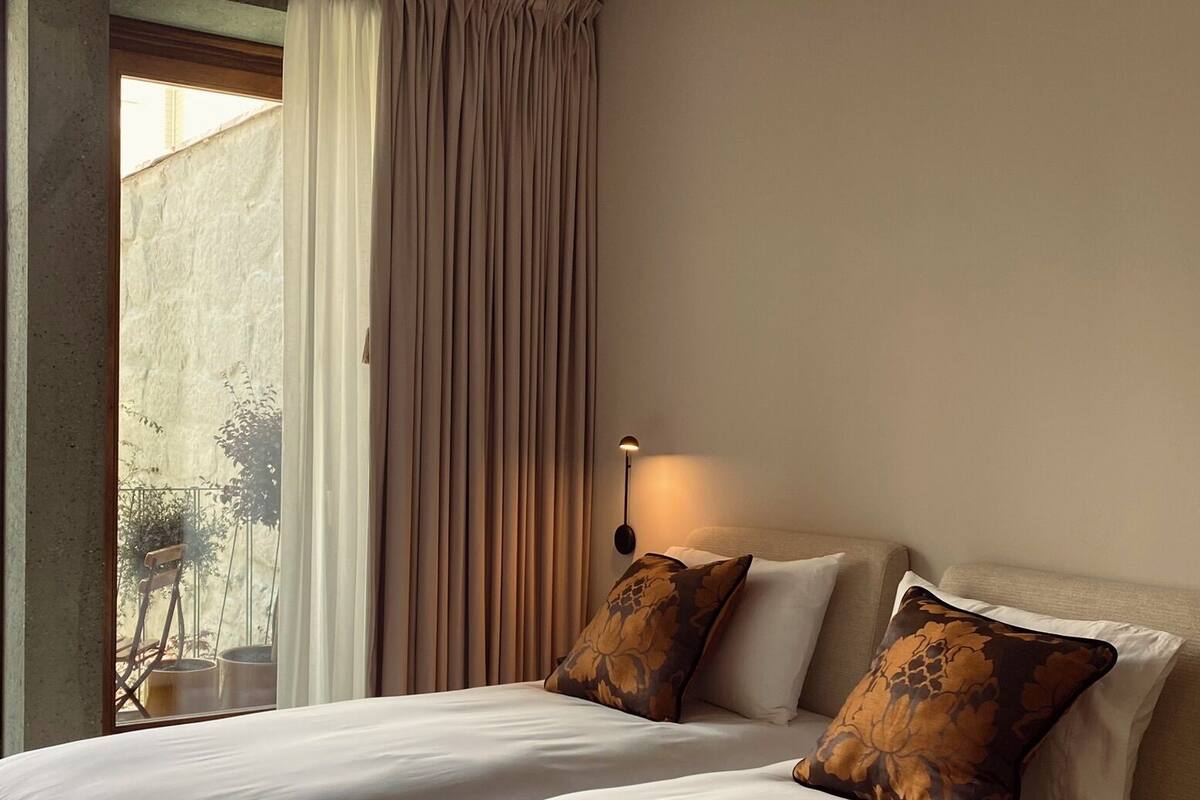 Una cama doble con sábanas blancas y dos almohadas decorativas, una lámpara de noche y vistas a un balcón con cortina.