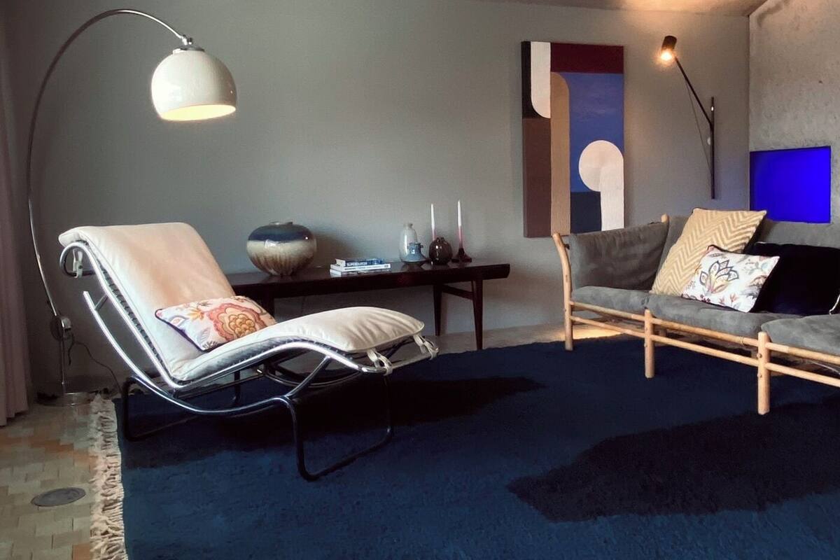Un dormitorio tipo loft moderno con techo alto, un ventanal grande, una alfombra azul oscuro, un sillón, un sofá y una mesita con velas.